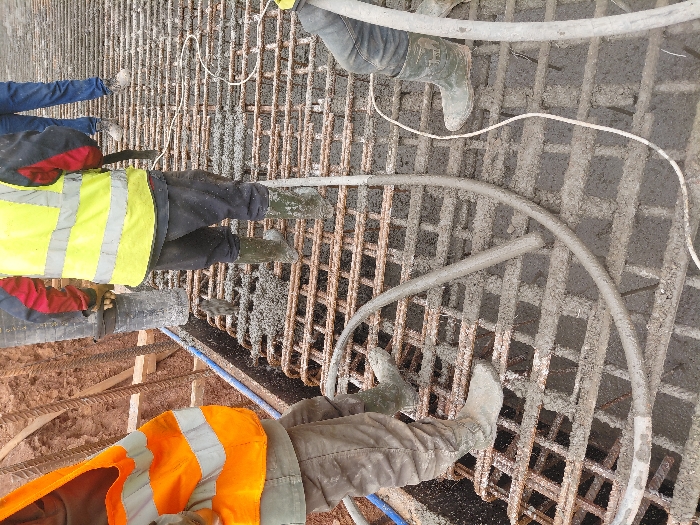 The scene depicts a concrete pouring operation at a construction site in Fdalate, Morocco. Three individuals are actively engaged in the process, with a fourth partially visible. The central worker, wearing a yellow high-visibility vest with reflective stripes over a red and black long-sleeved garment, dark trousers, and green rubber boots, is operating a concrete pump hose, directing wet concrete onto a steel rebar grid. This individual also holds a concrete vibrator, indicated by a black tool with an attached white cable. To the left, a worker in an orange high-visibility vest, dark trousers, and green rubber boots stands on the rebar, partially obscured. A third individual, wearing blue jeans and light-colored shoes, is positioned further back, partially visible behind the central worker. The lower leg and green rubber boot of a fourth worker are visible on the far right. The steel rebar grid is partially filled with fresh, grey concrete. A thick, grey concrete delivery hose, coated in concrete, is coiled on the grid, alongside a thinner white electrical cable. The background consists of exposed brown soil and wooden formwork components.