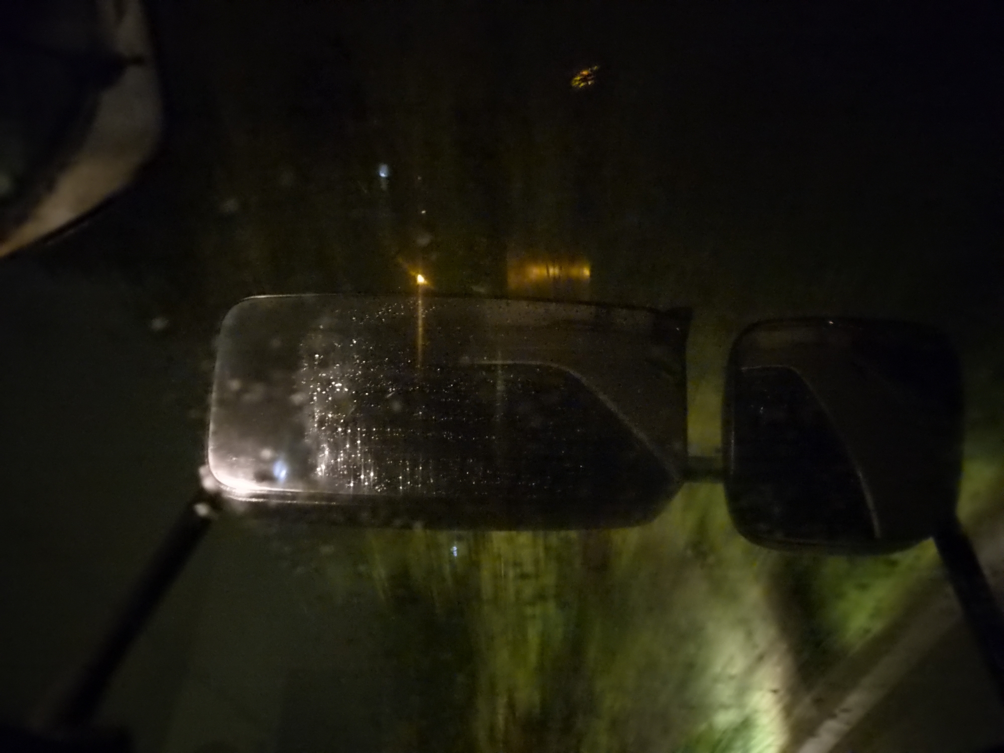 The image depicts a driver's-side view of a vehicle's side mirrors at night. Water droplets are visible on the window and mirror surfaces. Through the mirrors, a dark, indistinct environment is visible with sparse illumination, possibly reflecting vegetation or street lighting. The location is unidentifiable as Leudelange, Luxembourg, from the visual information alone. No people are visible in the image.