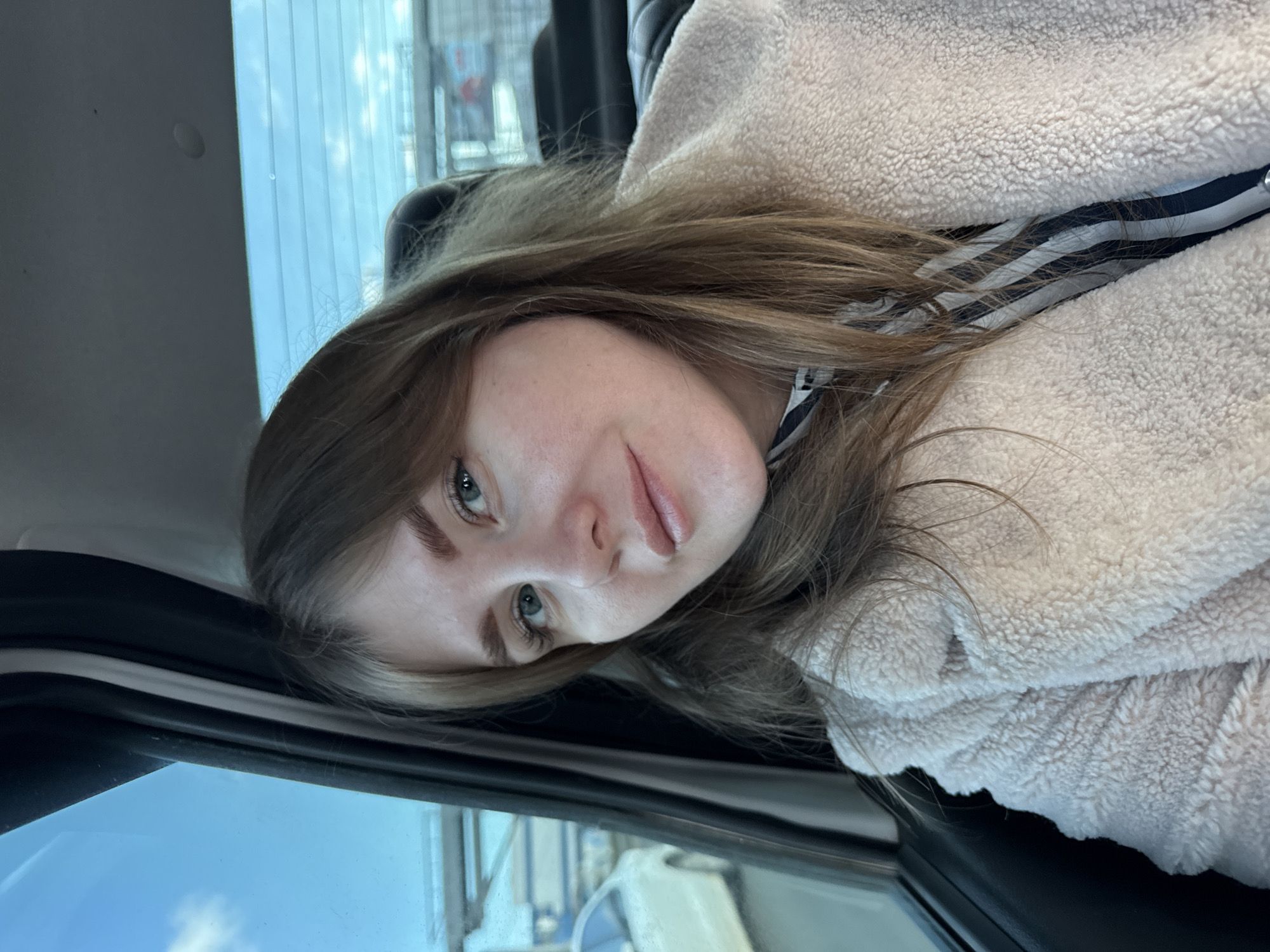 A young woman with light brown hair and fair skin is visible in the foreground of the image. She is wearing a light-colored, fuzzy jacket over a black and white striped garment. Her face is angled towards the camera, and her gaze is directed forward. The background shows a blurred urban environment with hints of buildings and a light blue sky visible through a window. The image appears to have been taken inside a vehicle, judging by the presence of the car's interior frame. It is unknown whether the location is the city of Perm, Russia.