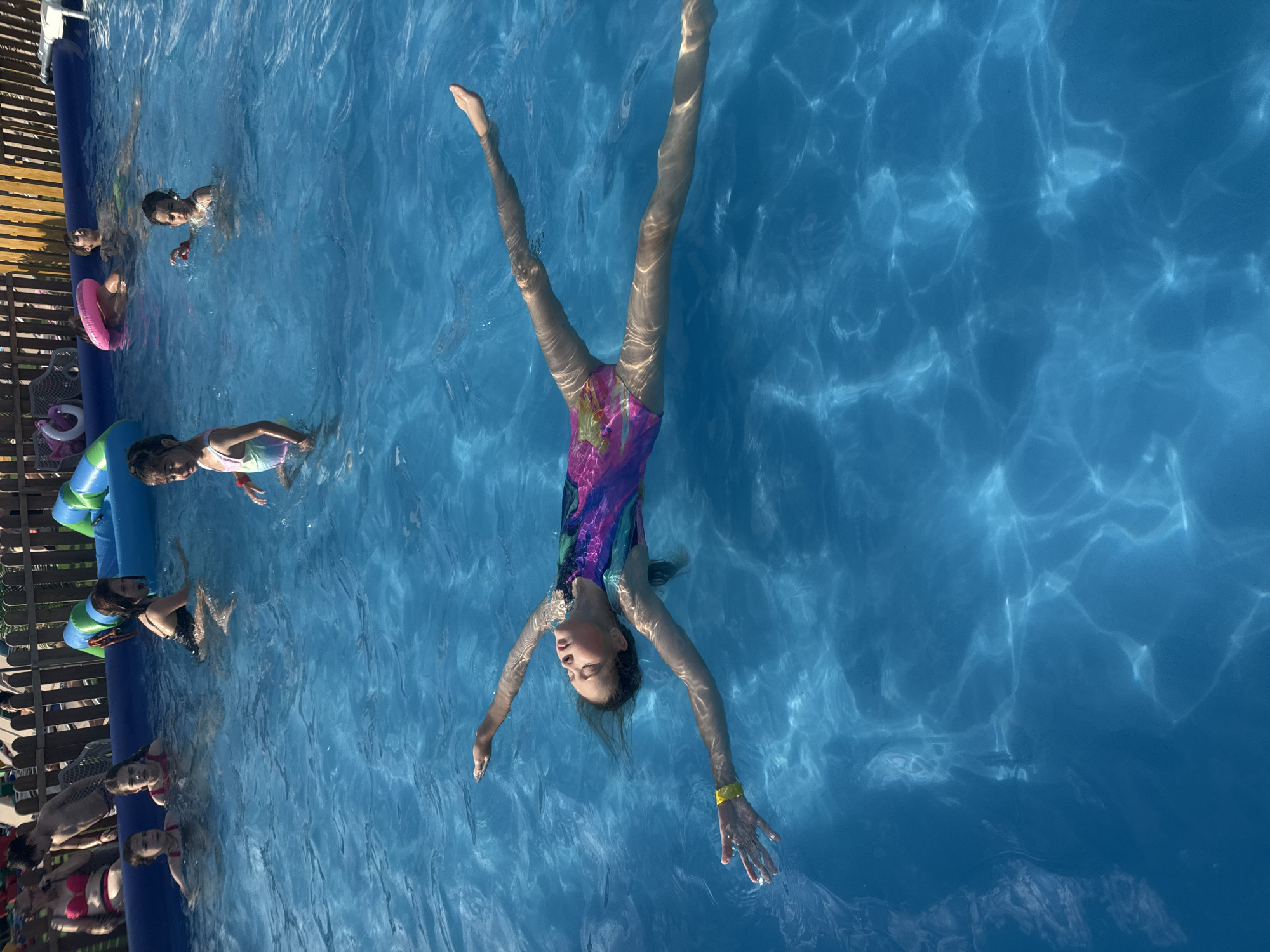 In an outdoor swimming pool, a young girl floats on her back, her eyes closed, appearing relaxed. She is wearing a colorful swimsuit and a yellow wristband. Sunlight refracts through the clear blue water, creating shimmering patterns on the pool's surface. Other children and adults are visible in the background, some in the water and others on the poolside, enjoying a sunny day. The setting appears to be a private backyard or a small community pool area, given the wooden fence and inflatable water toys. The location is identified as Rusanovo, Russia.