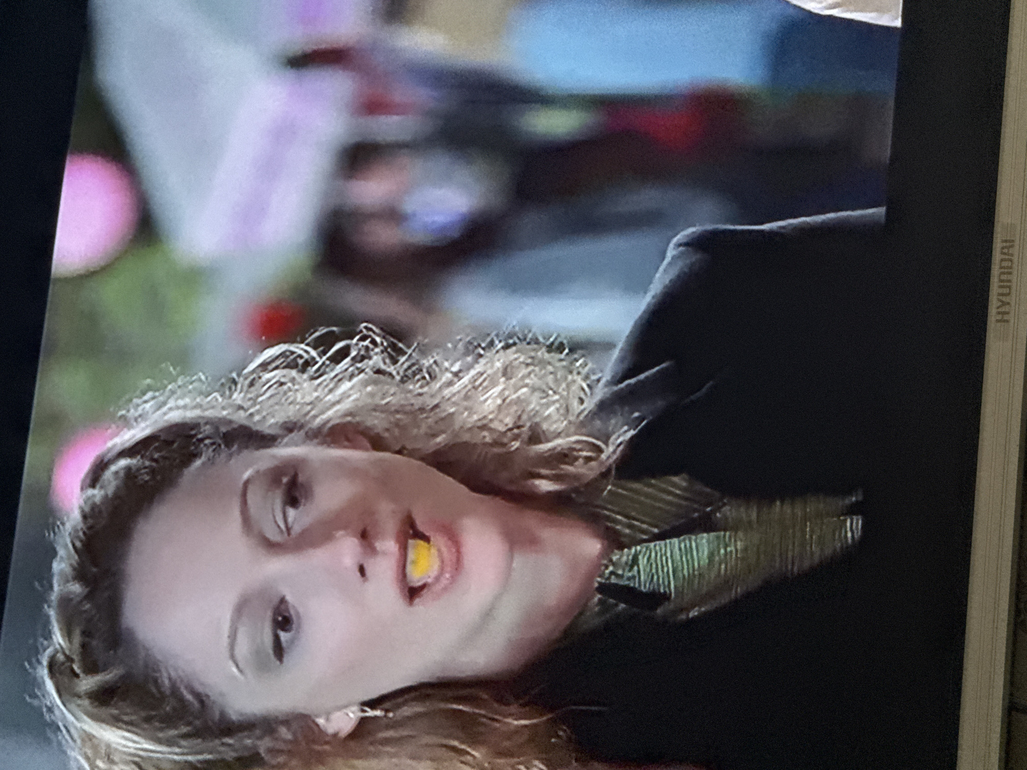A woman with curly hair is looking at the camera with a yellow object in her mouth. She is wearing a dark blazer over a gold and green striped shirt. The background is blurry but appears to show pink and white decorations, possibly balloons. The location cannot be identified as Mytishchi, Russia based on the information in this single image.