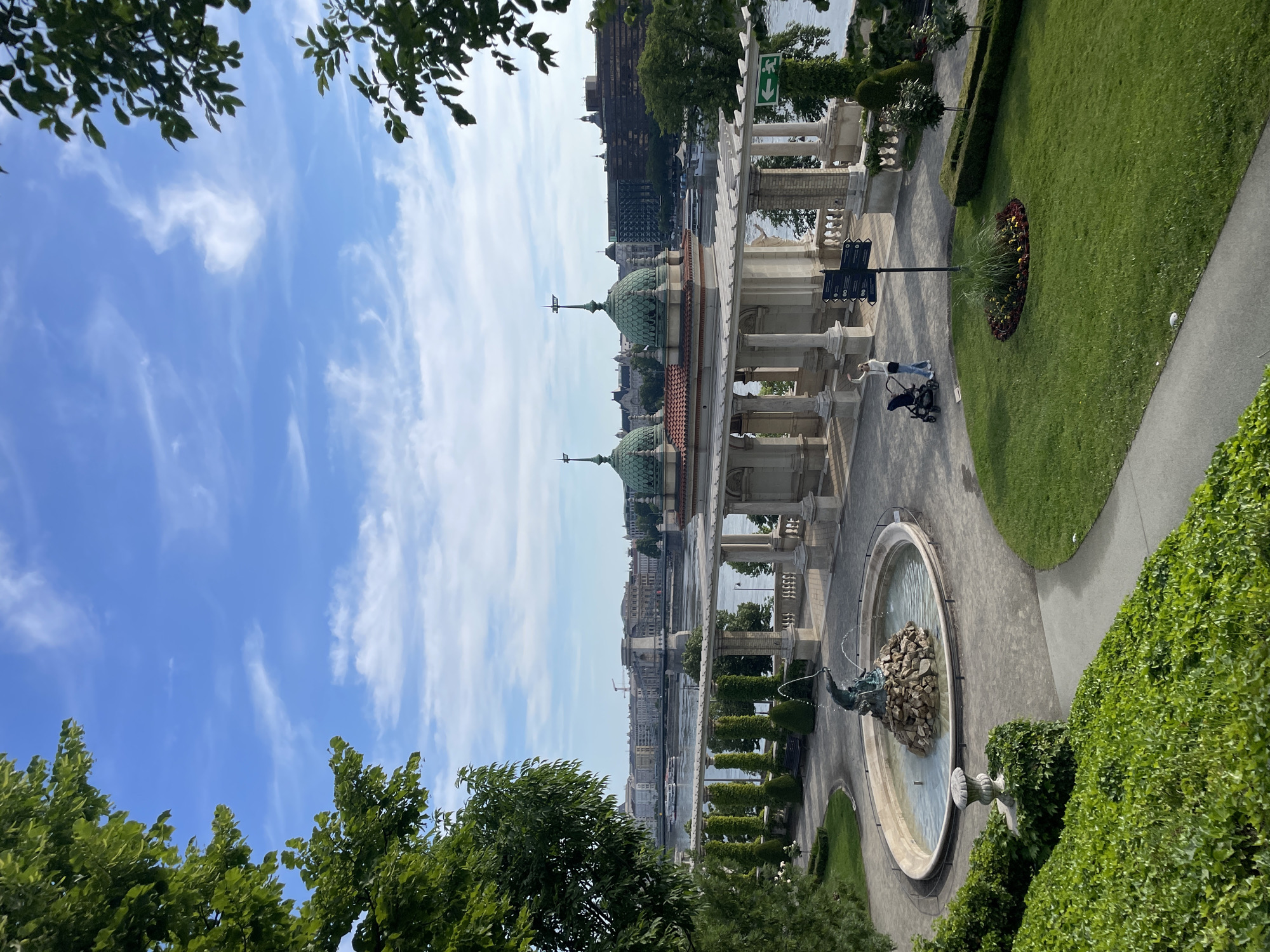 The image depicts a high-angle view of a cityscape in Budapest, Hungary. The scene is dominated by a complex of buildings with green domes and a colonnaded facade, suggesting a historical or public structure.
In the foreground, a circular fountain with a statue in the center is visible, surrounded by manicured green spaces and hedges. A path runs alongside the fountain, where a woman with a stroller appears to be walking.
Beyond the buildings, the Danube River is seen, with various buildings and structures lining its banks. The sky is a bright blue with wispy clouds. The overall impression is of a well-maintained public space within a historic urban setting.