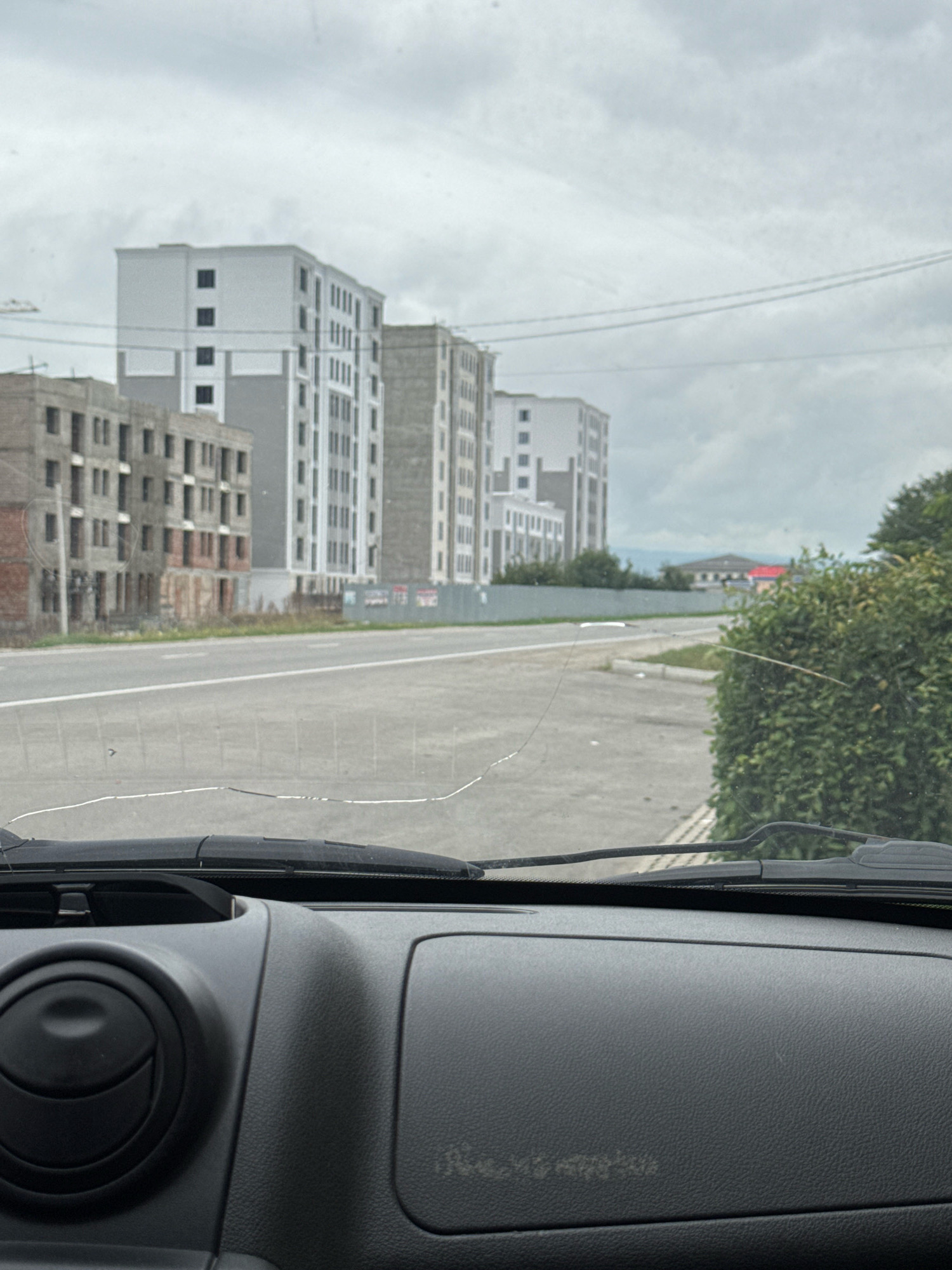 The image, taken from inside a vehicle, shows a view through the windshield of a street in Baksan, Russia. In the background, several modern apartment buildings are visible, some appearing to be under construction. A paved road with lane markings runs through the middle ground, bordered by a green hedge on the right. The sky is overcast with grey clouds. The foreground of the image shows the interior of the vehicle, including the dashboard, windshield wipers, and a portion of the driver's side air vent. A crack is visible on the windshield. There are no people or animals in the scene.
