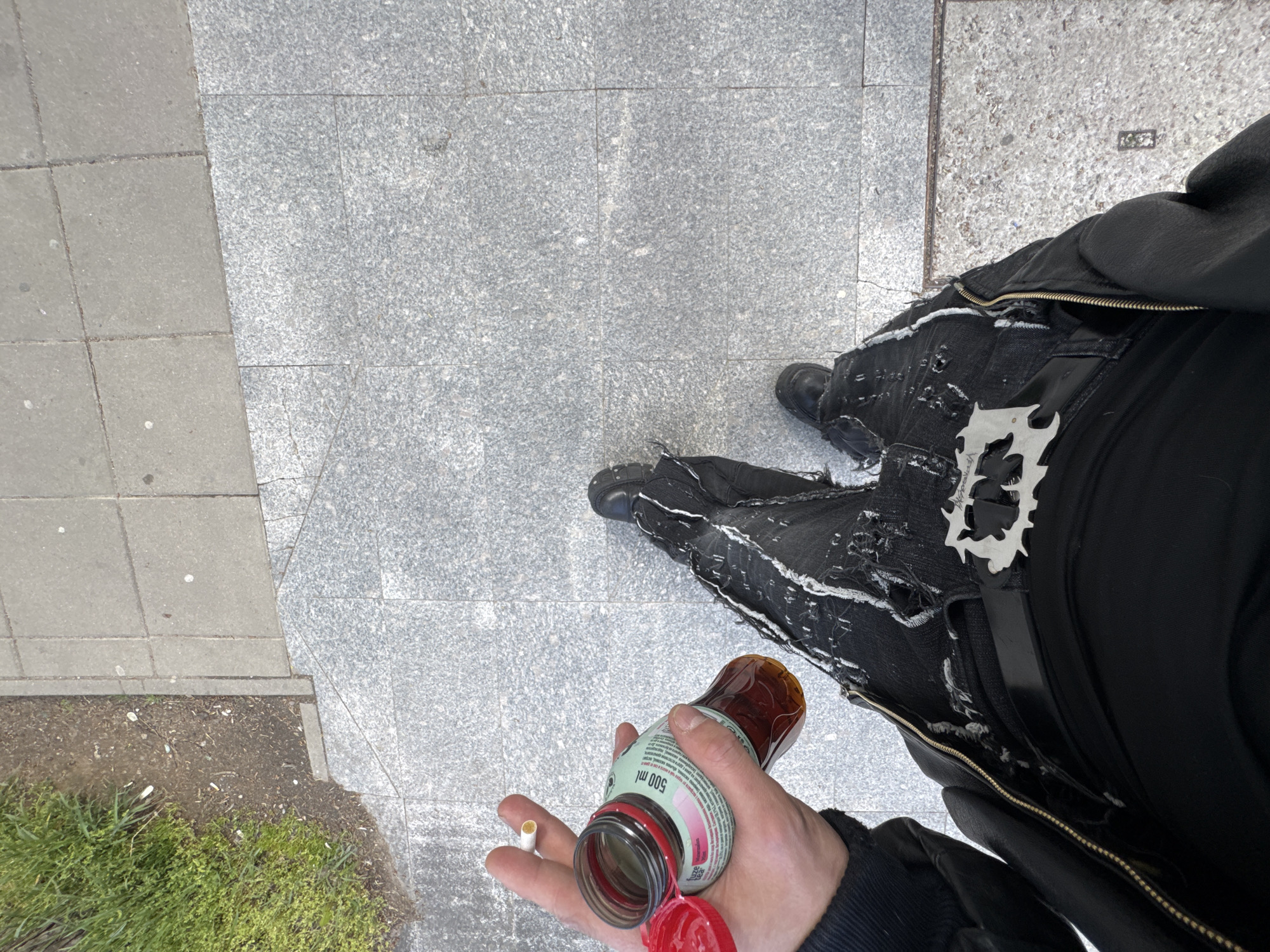 An overhead, high-angle shot captures a person standing on a paved surface in Sofia, Bulgaria. The image focuses on the person's lower body, dressed in heavily distressed black jeans with visible white threads and sturdy black boots. The person's right hand is prominent in the foreground, holding a plastic bottle with a bright red, partially open cap. The bottle, containing a dark reddish-brown liquid, has "500 ml" clearly visible on its label. A cigarette filter is held between the thumb and forefinger of the same hand. A metallic, abstractly shaped buckle or ornament is attached to the right hip area of the jeans.
The ground beneath the person is a mosaic of urban paving materials: light grey granite tiles with a speckled texture on the right, and darker grey, smoother concrete pavers on the left. These two surfaces meet with a distinct diagonal line. In the bottom left corner, a small patch of dirt and green ground cover is visible. The lighting is bright and even, indicating daytime conditions, possibly under an overcast sky. The image offers a glimpse of an individual's attire and a casual moment in an urban setting.