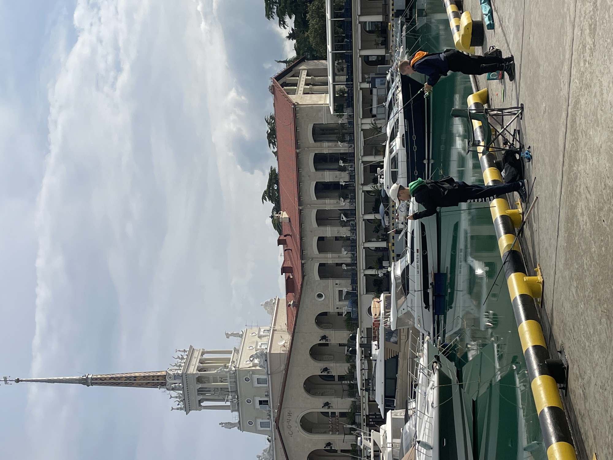 Two people are fishing from a dock in Sochi, Russia. The dock has black and yellow striped bumpers along the edge. Yachts are moored alongside the dock. A building with a tall spire and arched balconies is visible in the background. The sky is partly cloudy.