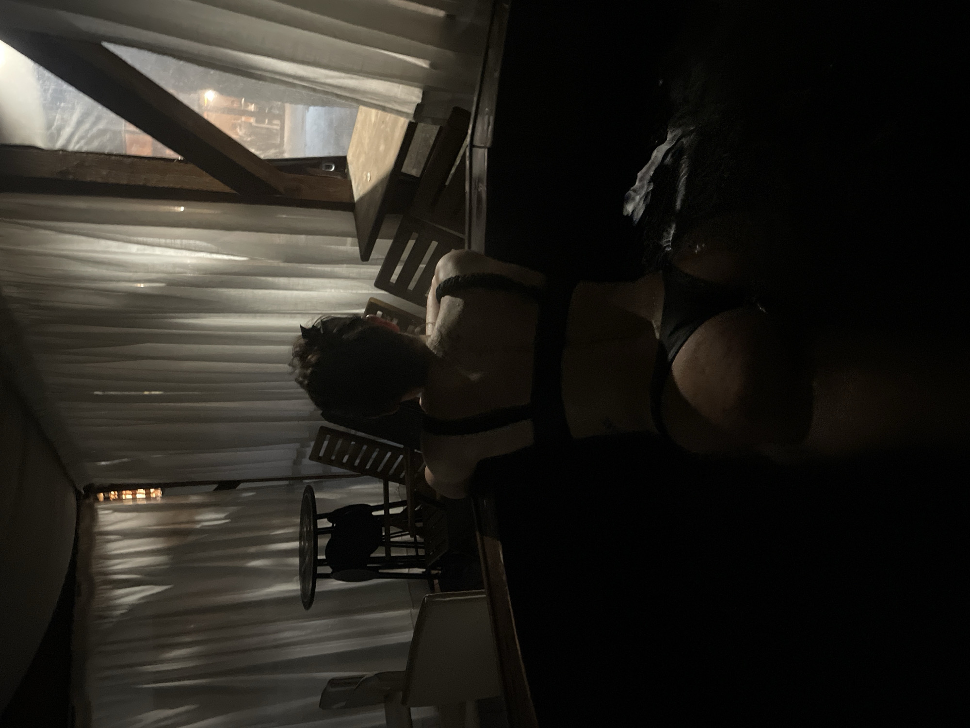 A person wearing minimal clothing, specifically a back-revealing garment and dark undergarments, is sitting or lying on a surface in a dimly lit indoor location. The location appears to be decorated with draped fabric or curtains, creating a soft, textured backdrop. The presence of chairs and a table suggests a possible indoor dining or lounge area. The person's hair is short and dark. The image is dark with the background details suggestive of an indoor space, with limited visibility. There is no verifiable evidence to suggest this is in Alino, Bulgaria.