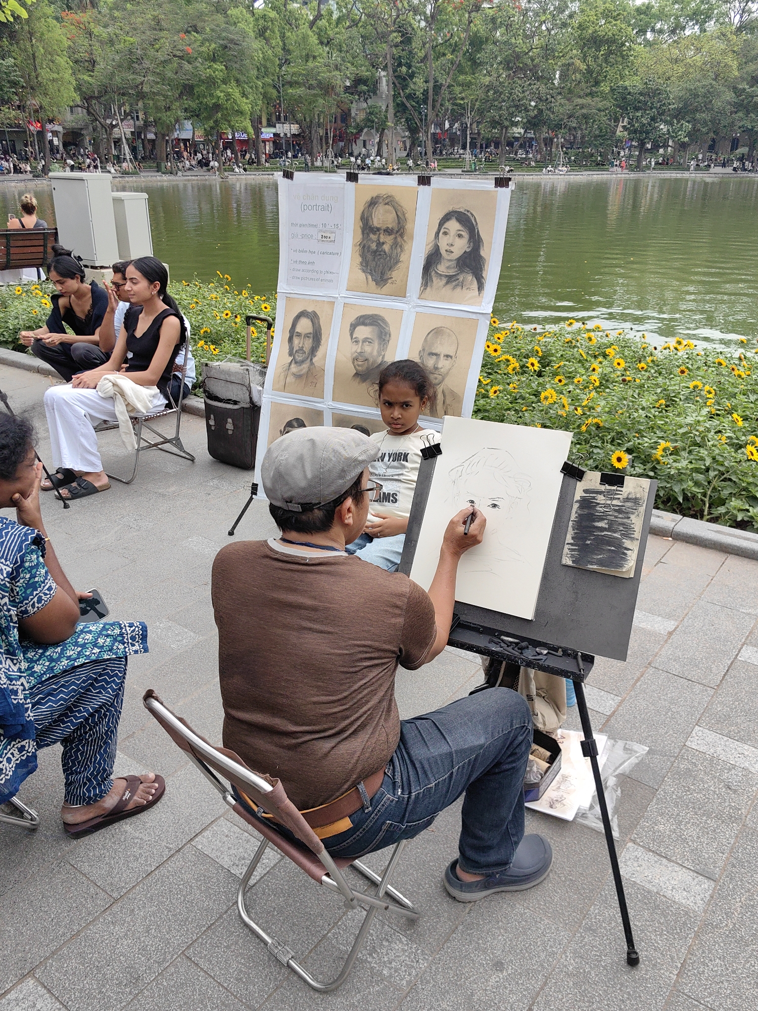 An outdoor scene captures a street artist creating a portrait by a tranquil lake, likely Hoan Kiem Lake in Hanoi, Vietnam. In the foreground, a male artist wearing a grey cap, brown t-shirt, and blue jeans sits on a folding chair, intently sketching on an easel. His subject, a young girl with dark hair, sits opposite him, looking towards the artist with a calm expression. She wears a white t-shirt with partial text "NEW YORK" and "AMS" visible.
Adjacent to the artist, a display board showcases various examples of his work, including charcoal portraits of a bearded man, a young girl, and several men resembling celebrities like Keanu Reeves and Jason Statham. The board also features a sign with text in Vietnamese and English: "vẽ chân dung (portrait)," indicating a drawing service. It specifies "thời gian (time): 10-15" (minutes) and "giá-price: 200" (likely 200,000 VND). Other services advertised include "* về biểm họa (caricature)," "* về theo ảnh (draw according to photo)," and "draw pictures of animals." Unclear text "SNA" and "GXUE" is visible on background signs.
Several people are present in the midground, observing or waiting. To the left, a woman in a blue and white patterned outfit sits on a chair, while two other women are also seated, one in white pants and a black top, and another in a black top.
Behind the artist and display, a vibrant bed of yellow sunflowers lines the edge of a serene green lake. The opposite bank of the lake features a tree-lined promenade dotted with numerous people and buildings under bright daylight, suggesting a lively urban park setting. The overall atmosphere is one of calm creativity amidst a bustling public space.