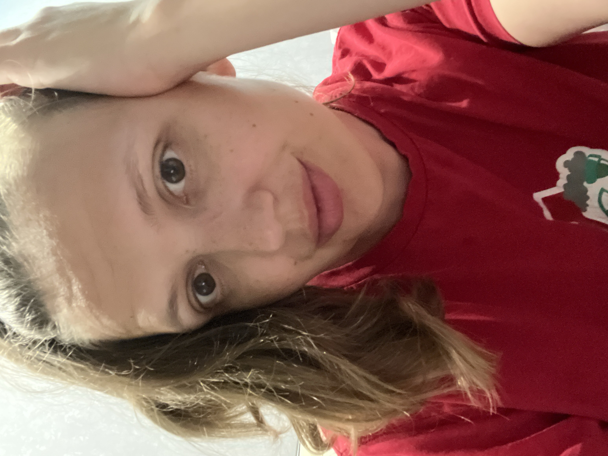A woman with fair skin and brown eyes looks at the camera. She has freckles on her face. Her hair is blonde and brown. She is wearing a red shirt with a design on the left side of the chest. Her left hand is resting on her forehead. The background appears to be a white surface. There is no information in the image to indicate that this image was taken in Chaikovsky, Russia.