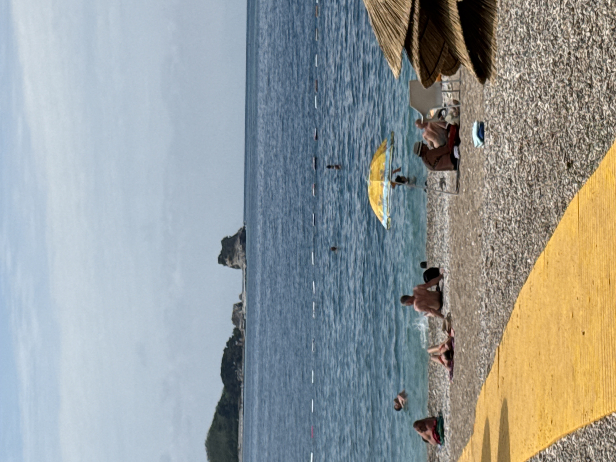 The image depicts a vibrant daytime scene at a pebbly beach in Budva, Montenegro. The calm, blue sea stretches into the distance, marked by a line of white buoys, with a rocky, vegetated coastline visible on the horizon to the left. The sky above is a pale blue with subtle cloud formations.
Numerous individuals are enjoying the beach and water. Several people are swimming or wading in the sea, including one person fully submerged further out and another standing closer to the shore. A distinctive yellow umbrella or large flotation device is visible floating in the water.
On the pebbly shore, a few individuals are relaxing. One person is seen lying down near the water's edge. Further back, under the shade of a large straw beach umbrella (partially visible in the upper right), two people are seated on lounge chairs. A blue towel or piece of clothing is also visible on the gravel.
A prominent feature in the foreground and along the right side of the image is a bright yellow, textured walkway or path that curves along the beach, separating it from an unseen area to the right. The overall atmosphere suggests a leisurely summer day, with people enjoying the sun and sea. There is no visible text in the image.