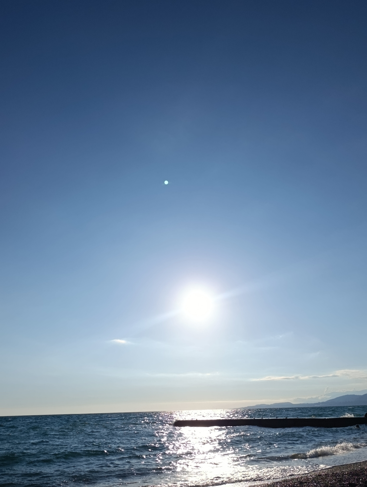 This media file depicts a serene seascape with a clear blue sky. The sun is prominently featured, shining brightly and casting a shimmering reflection on the rippling water. A dark, rectangular structure, possibly a pier or breakwater, extends into the sea from the right side of the frame. The waves are gently lapping at a pebble beach in the foreground. In the distance, hazy mountains or hills can be seen against the horizon. The overall impression is one of a peaceful and sunny day at the coast. The bright sunlight suggests it is midday or late afternoon. The location is Eshyra, Abkhazia. There is no visible text in the image.