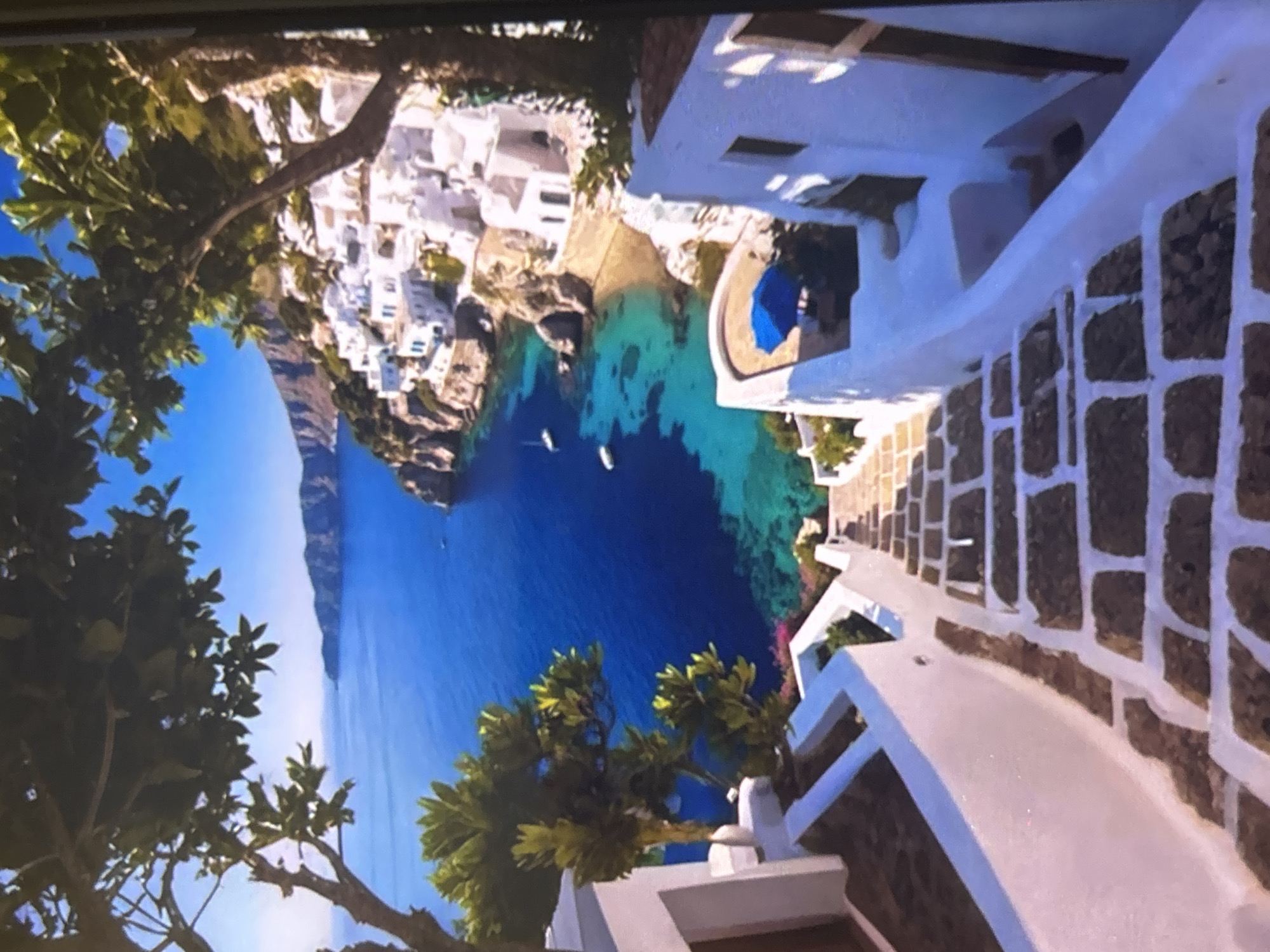 The media file is a vibrant, high-angle photograph of a picturesque coastal village, depicted as if viewed from an elevated vantage point on a sunny day. The image is currently rotated 90 degrees clockwise, but shows a landscape view when correctly oriented.
The scene features a stunning bay with exceptionally clear, multi-toned water transitioning from turquoise near the shore to deep blue further out. Several small boats are visible floating on the calm water. The bay is framed by a steep coastline with dramatic cliffs in the distance.
Clustered along the shore and ascending the hillside are numerous white-washed buildings, characteristic of traditional Mediterranean architecture, possibly from a Greek island. Lush green trees and foliage are prominent throughout the image, appearing in the foreground, mid-ground, and around the buildings. A winding pathway or set of stairs, constructed with stone and white mortar, leads down through white-walled structures on the right side of the image (when correctly oriented). A bright blue umbrella is visible near one of the buildings overlooking the water, suggesting a leisure area.
No people or specific activities are depicted, giving the scene a serene and tranquil atmosphere. The bright, clear blue sky indicates excellent weather, likely midday or early afternoon, with strong sunlight illuminating the landscape. There is no visible text in the image.