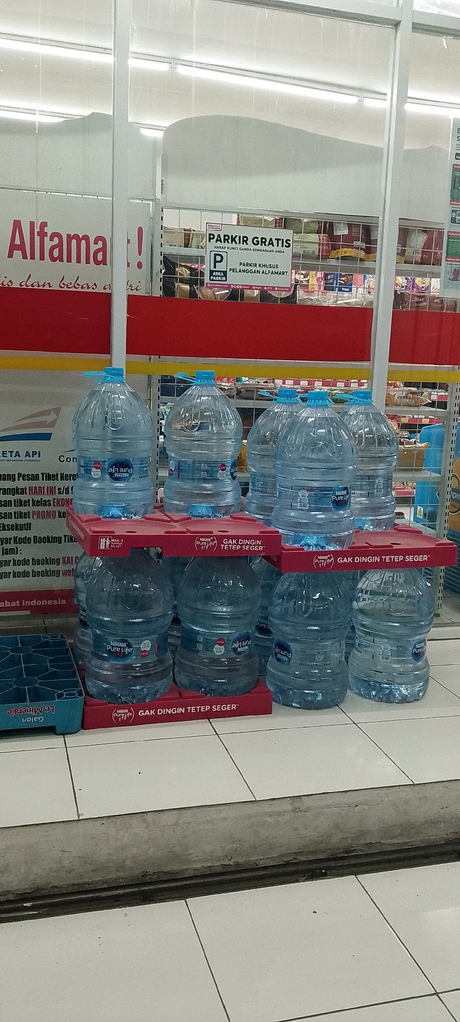In a retail store interior, likely a minimarket such as Alfamart, in Satriamekar, Indonesia, large-format plastic water bottles are displayed for sale. The bottles, filled with clear liquid, are arranged in tiered shelving units. Some bottles feature labels from brands like "Nestle Pure Life" and "Afnand". The shelving units are red and prominently display text such as "GAK DINGIN TETEP SEGER", which translates to "Not Cold, Still Fresh", a marketing slogan for the beverages. In the background, store signage is visible, including a parking sign for "PARKIR KHUSUS PELANGGAN ALFAMART" (Parking for Alfamart Customers). The floor is tiled in a grid pattern. The overall scene depicts a commercial environment focused on the sale of bottled water.
