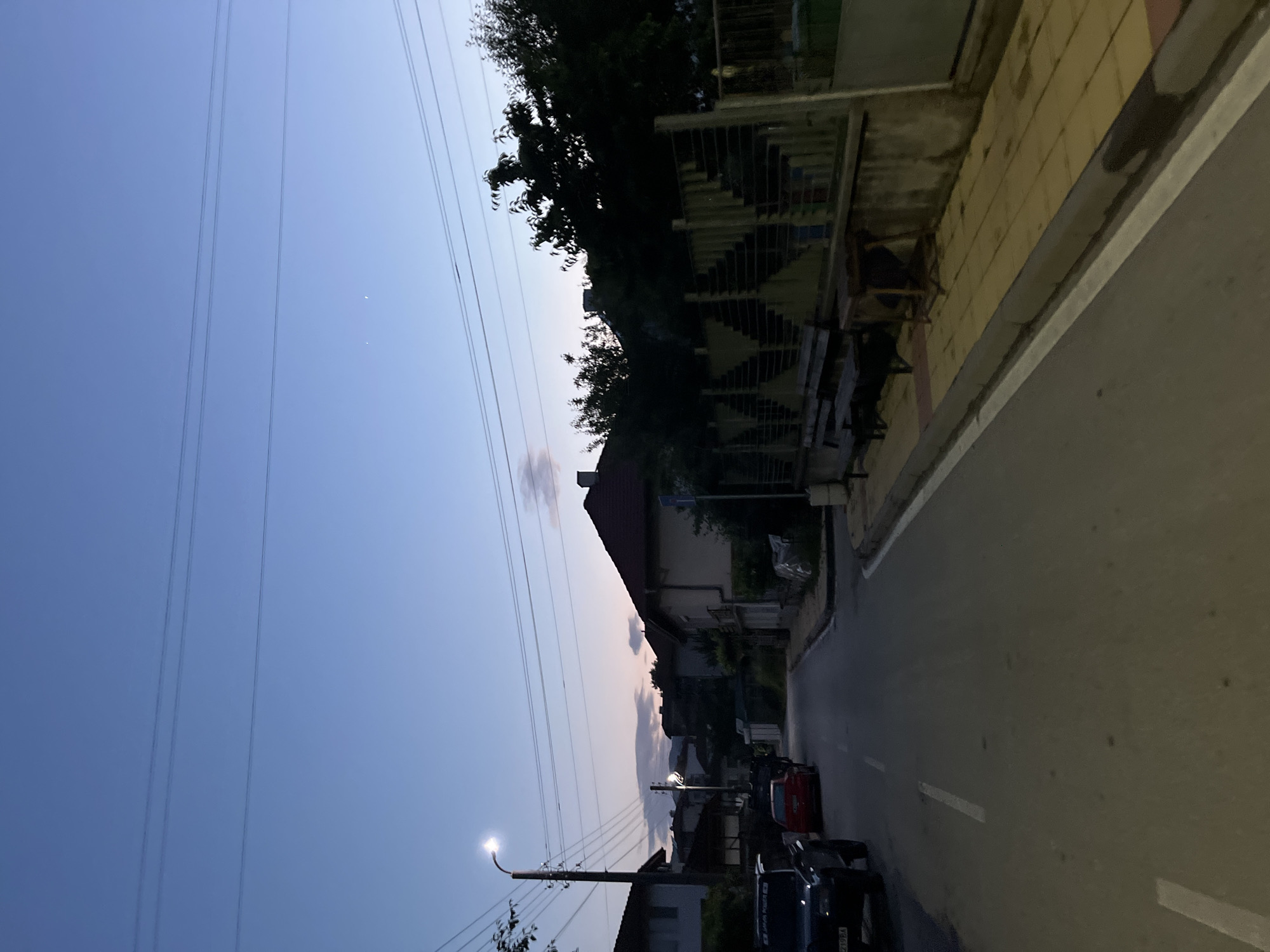 The image depicts a quiet residential street in Zlatograd, Smolyan Province, Bulgaria, during twilight. The scene is illuminated by an active streetlight on the left and the fading light of the sky.
A paved asphalt road with a dashed white centerline occupies the foreground and recedes into the distance. A solid white line delineates the curb on the right side of the road. On the left side of the street, a dark-colored SUV is parked, facing away from the camera. Its rear license plate, "X 2198 BA," is visible, identifying it as registered in the Smolyan region. Behind the SUV, a red car is partially visible, either parked or slowly moving. A tall, dark streetlamp with a bright, illuminated LED fixture stands prominently on the left, near the SUV. Multiple power lines run horizontally across the upper portion of the frame, above the street.
On the right side of the street, a sidewalk constructed of yellow/beige pavers with a red paver border is present. A wooden bench and a small, rustic wooden table are situated on the sidewalk, adjacent to a metal fence with a distinct zigzag or stepped geometric pattern. Behind this fence, dense trees and foliage are visible, casting deep shadows due to the low light. Houses with tiled roofs are discernible on both sides of the street further down the road.
The sky exhibits a twilight gradient, transitioning from a deep, clear blue overhead to a lighter, warmer hue of pale orange and pink near the horizon, where a few faint clouds are scattered. Two small, bright celestial objects (likely stars or planets) are visible in the upper-right quadrant of the sky. No individuals are visible in the scene.
