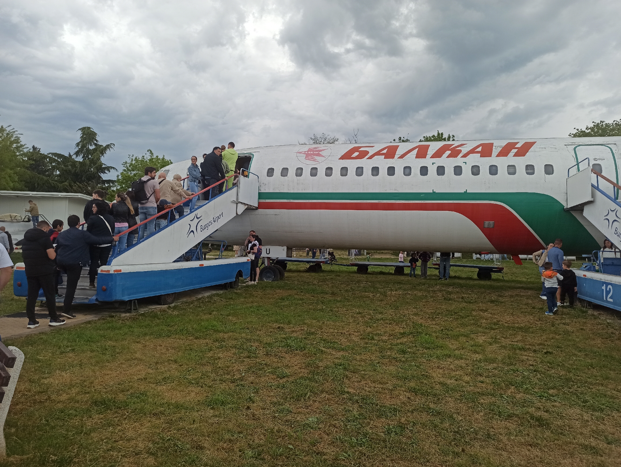 The image shows an old "Balkan" aircraft in Burgas, Bulgaria. A group of people are entering and exiting the plane via mobile stairs. The aircraft is white with a red "БАЛКАН" logo and a green and red stripe. The stairs have the text "Burgas Airport" printed on them. The background shows an overcast sky and sparse vegetation.