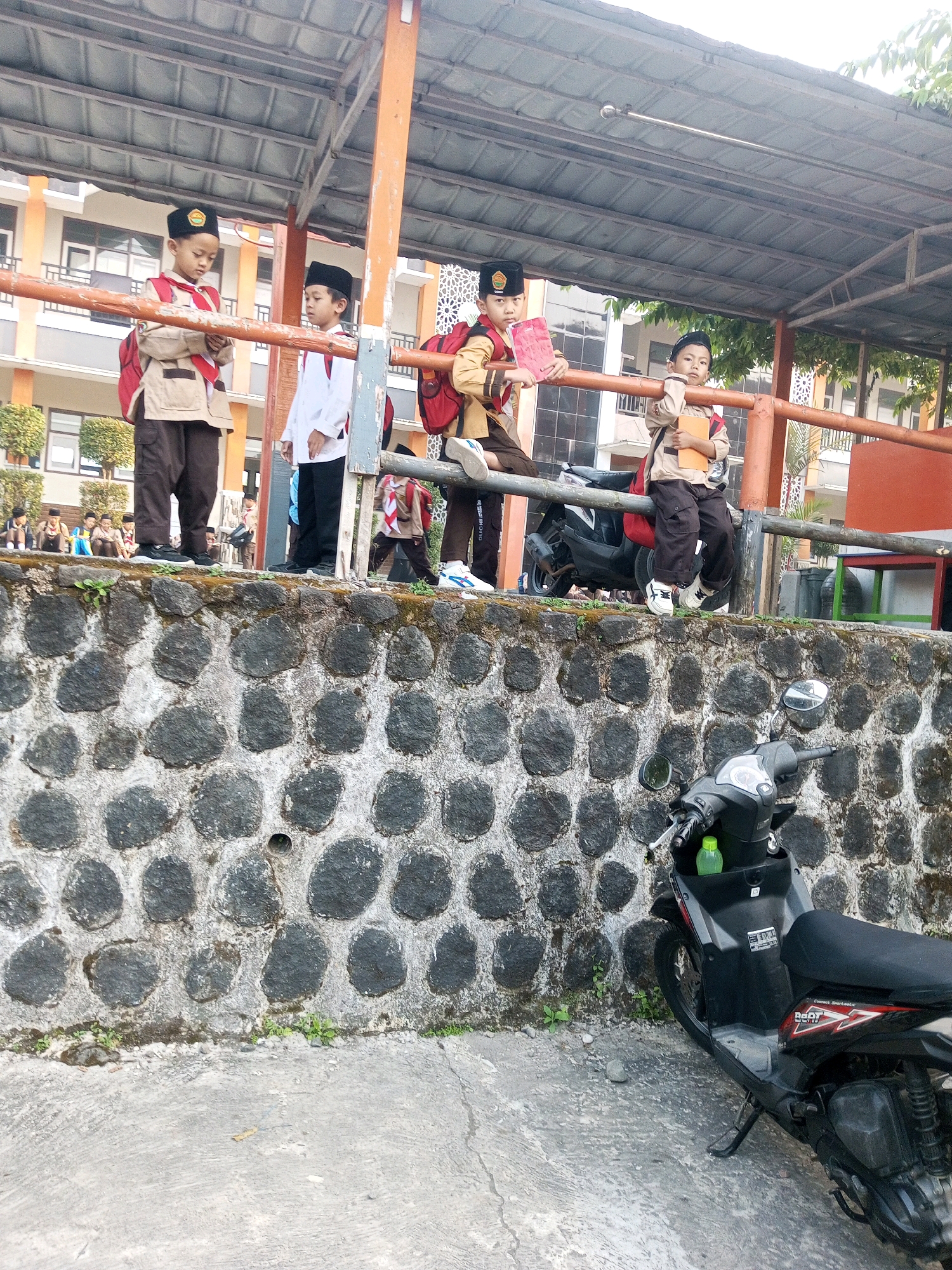 Four young boys in school uniforms are standing on a raised platform near a stone wall. The boys are wearing dark caps, beige shirts, and dark pants. Two of the boys are holding books. A black scooter is parked on the concrete ground in front of the stone wall. In the background, a building with orange accents and windows is visible. The scene appears to be outdoors, possibly at a school in Krasak, Indonesia, as suggested by the context. The sky is overcast, and the lighting suggests it is daytime.