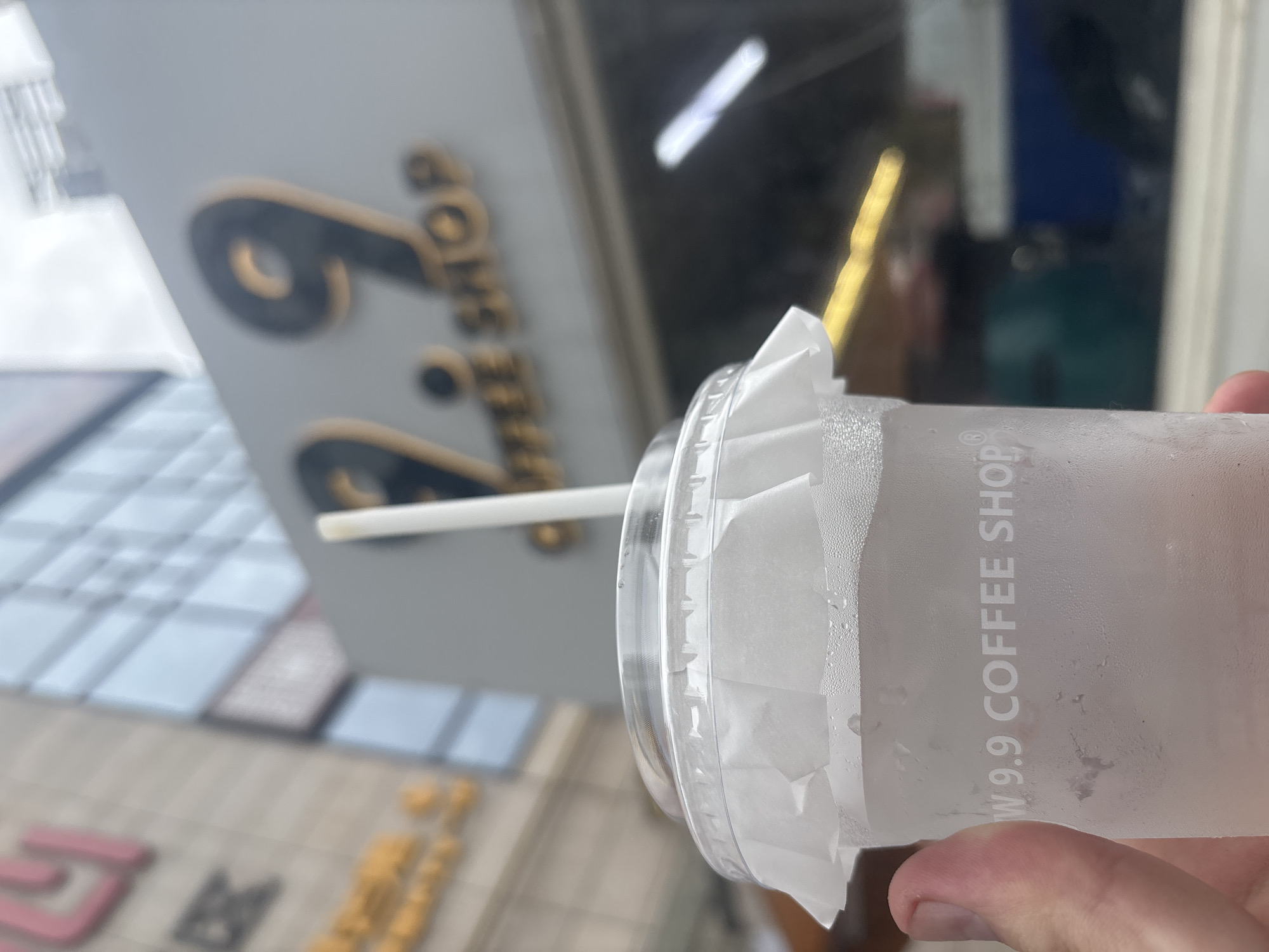 An outdoor shot focuses on a translucent plastic cup of iced beverage. A hand grips the cup from the left. Condensation forms on the cup's exterior. A white paper sleeve is folded around the top of the cup, and a white straw is inserted into the cup. In the background, a sign with the numbers "9.9" and "COFFEE SHOP" in large, light brown lettering is visible. The sign is mounted on a gray facade. Parts of a building with reflective windows are visible behind and to the left of the sign. The overall setting appears to be an urban environment in Nanhai District, China.