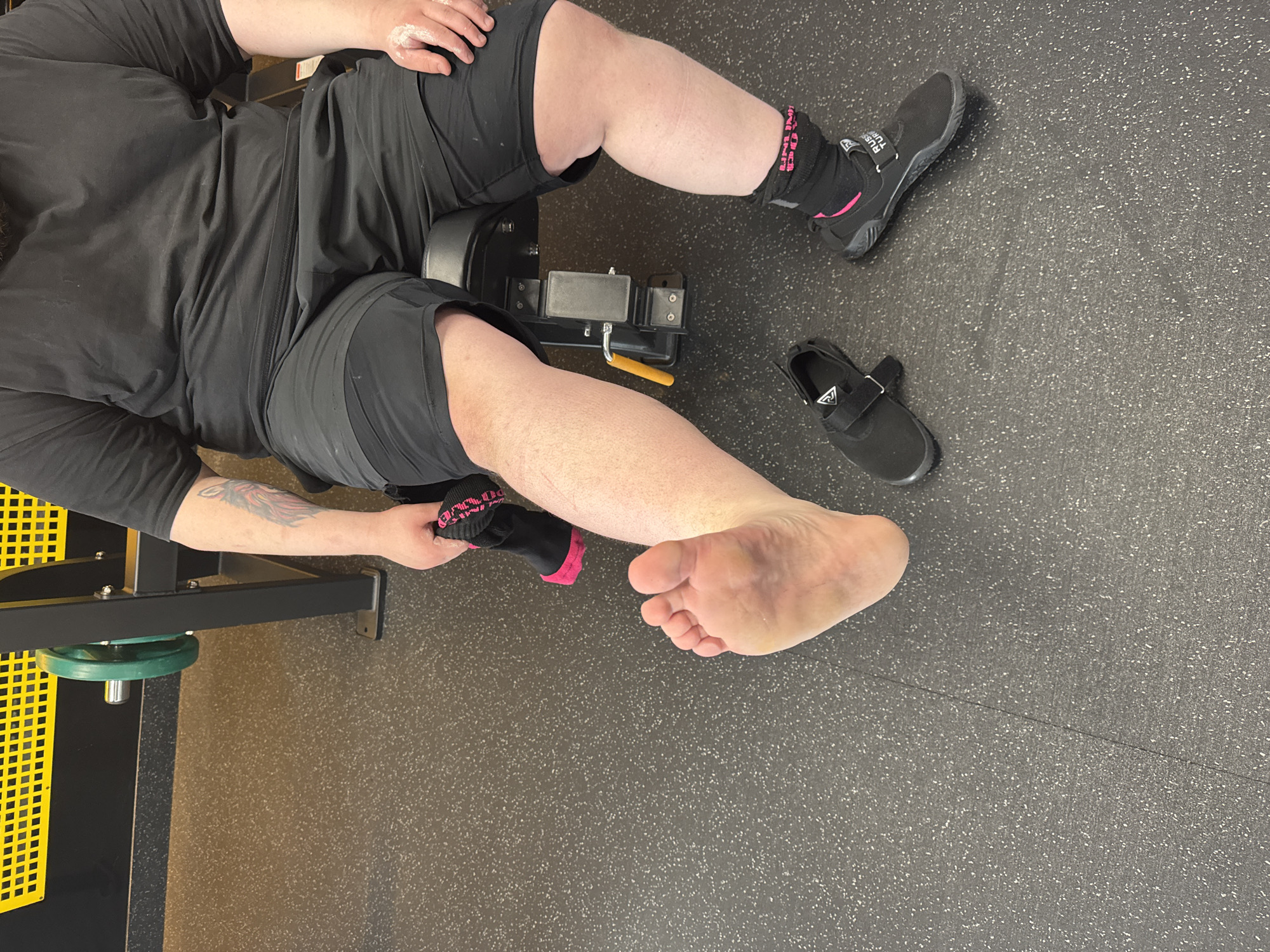 An overhead, medium shot captures a single, unidentified individual seated within an indoor gym setting. The person, appearing to be an adult male with a heavy build, wears a black short-sleeved t-shirt and black shorts over black compression shorts. A partial view of a tattoo, depicting a winged creature, is visible on the upper left arm. White chalk or powder residue is present on the person's right hand.
The individual is seated on a black upholstered gym bench. The left leg is extended forward, with the bare sole of the foot prominently displayed towards the viewer. The person holds a black ankle sock with pink accents in their left hand. A black minimalist-style shoe with a hook-and-loop strap rests on the dark gray speckled rubber gym floor near the bare left foot. The right foot, positioned closer to the bench, is wearing a similar black shoe, with a visible black and pink ankle sock underneath that displays "RUS TURF" on its strap.
In the background, elements of gym equipment are visible, including the dark metal frame of a machine with a green circular weight plate, and a yellow, perforated metal panel against a black wall. The floor throughout the scene is a dark gray, speckled rubberized material.