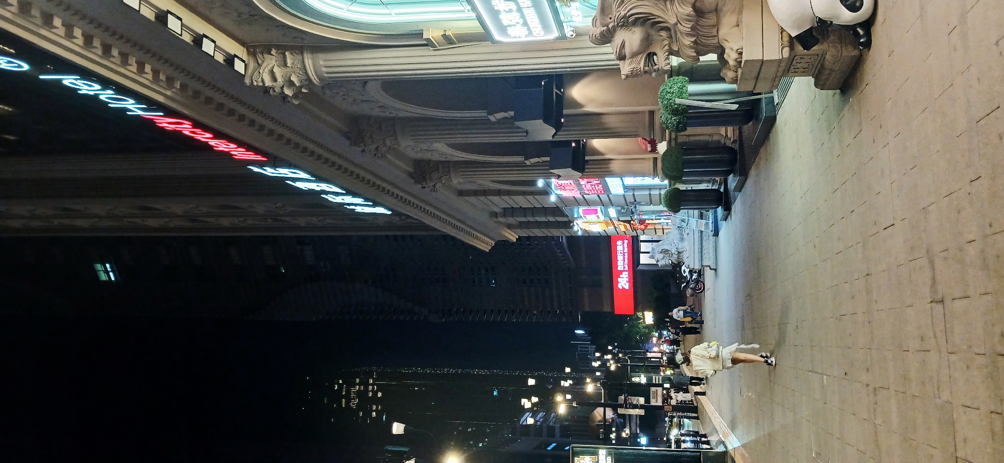 At night, the Intercity Hotel stands on Jiefangbei Pedestrian Street in Yuzhong District, Chongqing, China. The hotel exterior includes neon signs, stone columns, and lion statues. People stroll along the tiled walkway. Streetlights illuminate the scene. Buildings and city lights form the backdrop in the darkness.
