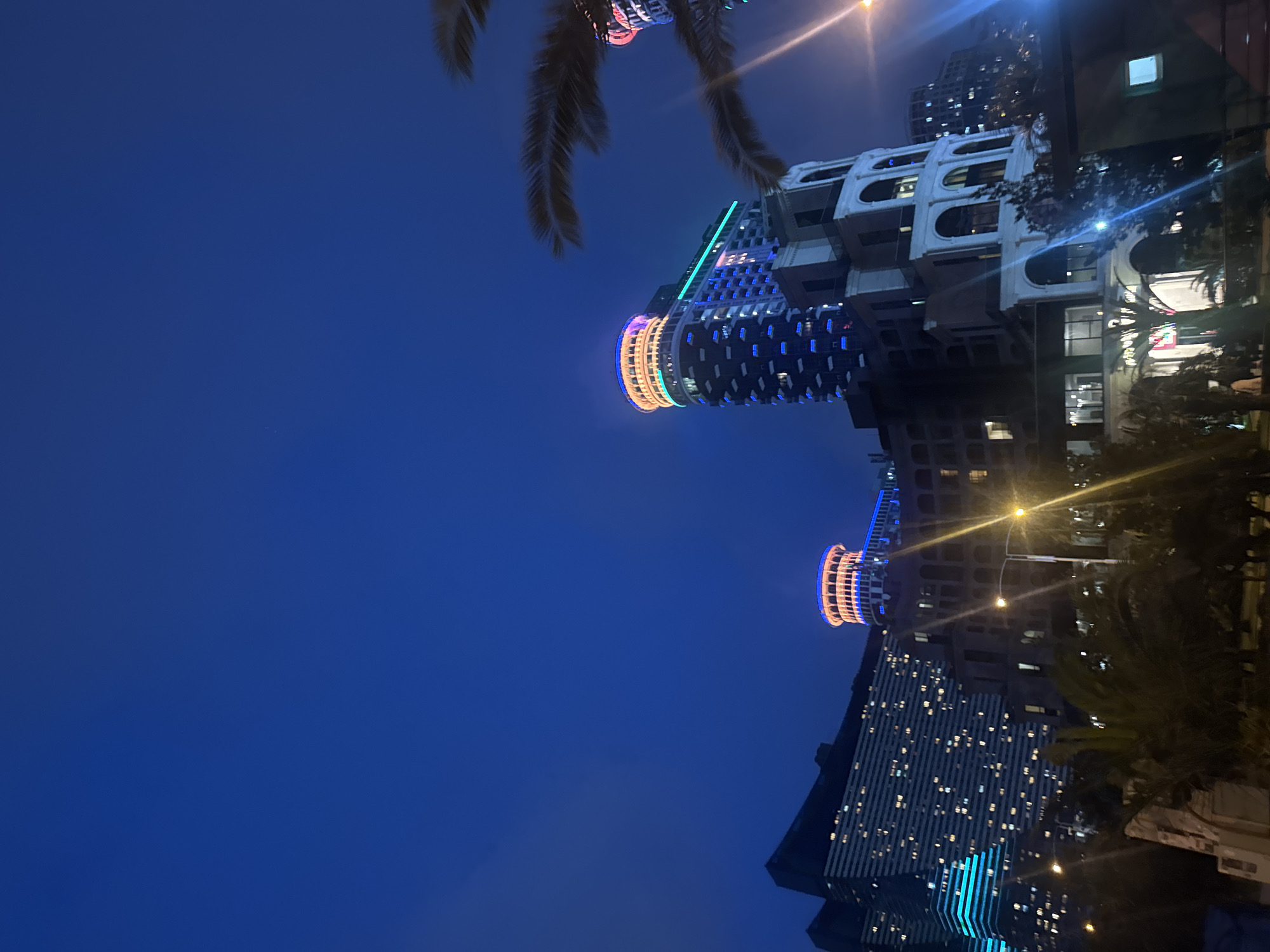 Here is a detailed description of the image:
The image shows a night scene in what appears to be a city, potentially near a coastal area. The most prominent feature is the modern architecture, including several high-rise buildings lit up with various colors of lights.
One building appears to have a cylindrical top with bands of colored lights. Another building features what looks like an arched, white facade. In the foreground, there are silhouetted palm tree fronds against a dark blue evening sky.
Streetlights are visible and illuminate some of the buildings and foliage. The overall lighting suggests it is either dusk or later in the evening. The scene appears to be a mix of residential and commercial areas, as indicated by the different types of buildings and their lighting.