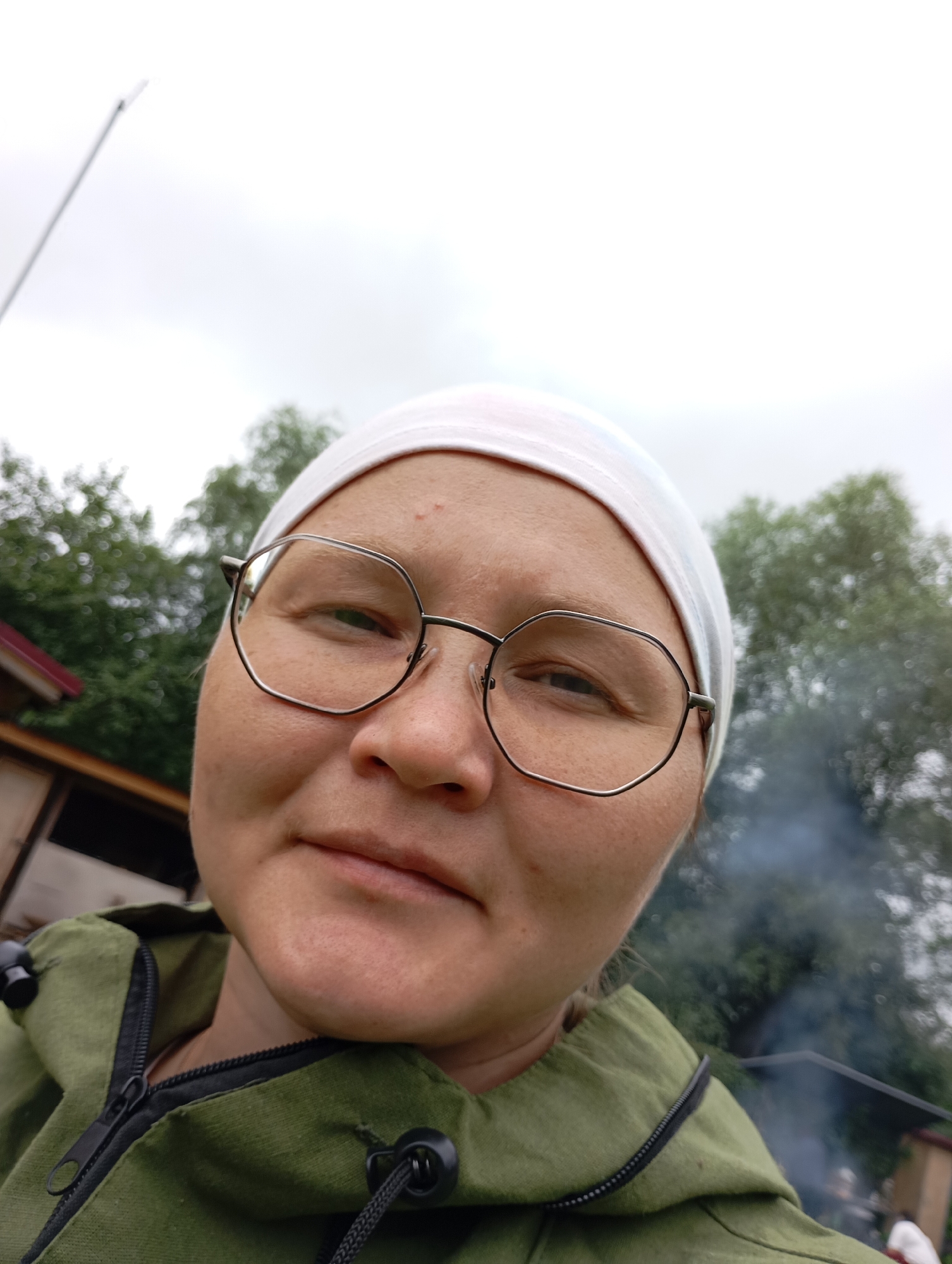 In the city of Malyi Bukor, Russia, a close-up, slightly upward-angled selfie shows a person wearing a white headscarf and modern, octagonal-framed glasses. They have fair skin with a few blemishes on their forehead. The person is wearing a green hooded jacket with a zipper. In the background, out of focus, are trees and what appears to be a building with a red roof, suggesting an outdoor setting, possibly a rural or suburban area. There is also a faint plume of smoke rising in the background, which could indicate a fire or cooking activity. The sky is overcast.