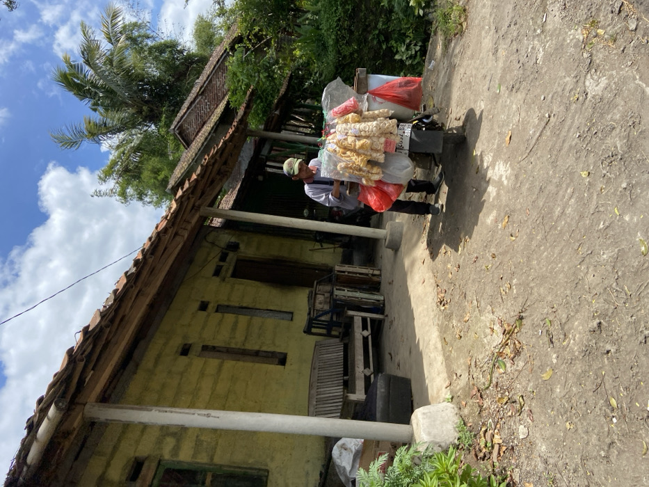 The image shows a man in a white shirt and a cap standing beside a motorcycle loaded with bags of food, located in front of a yellow house. The house has a wooden porch and a wooden bench. The motorcycle is parked on a dirt road. The man is facing the motorcycle and appears to be arranging the bags. There are trees and vegetation in the background. The scene is set in the village of Cipatat in Indonesia, at the coordinates -6.787 latitude and 107.434 longitude.