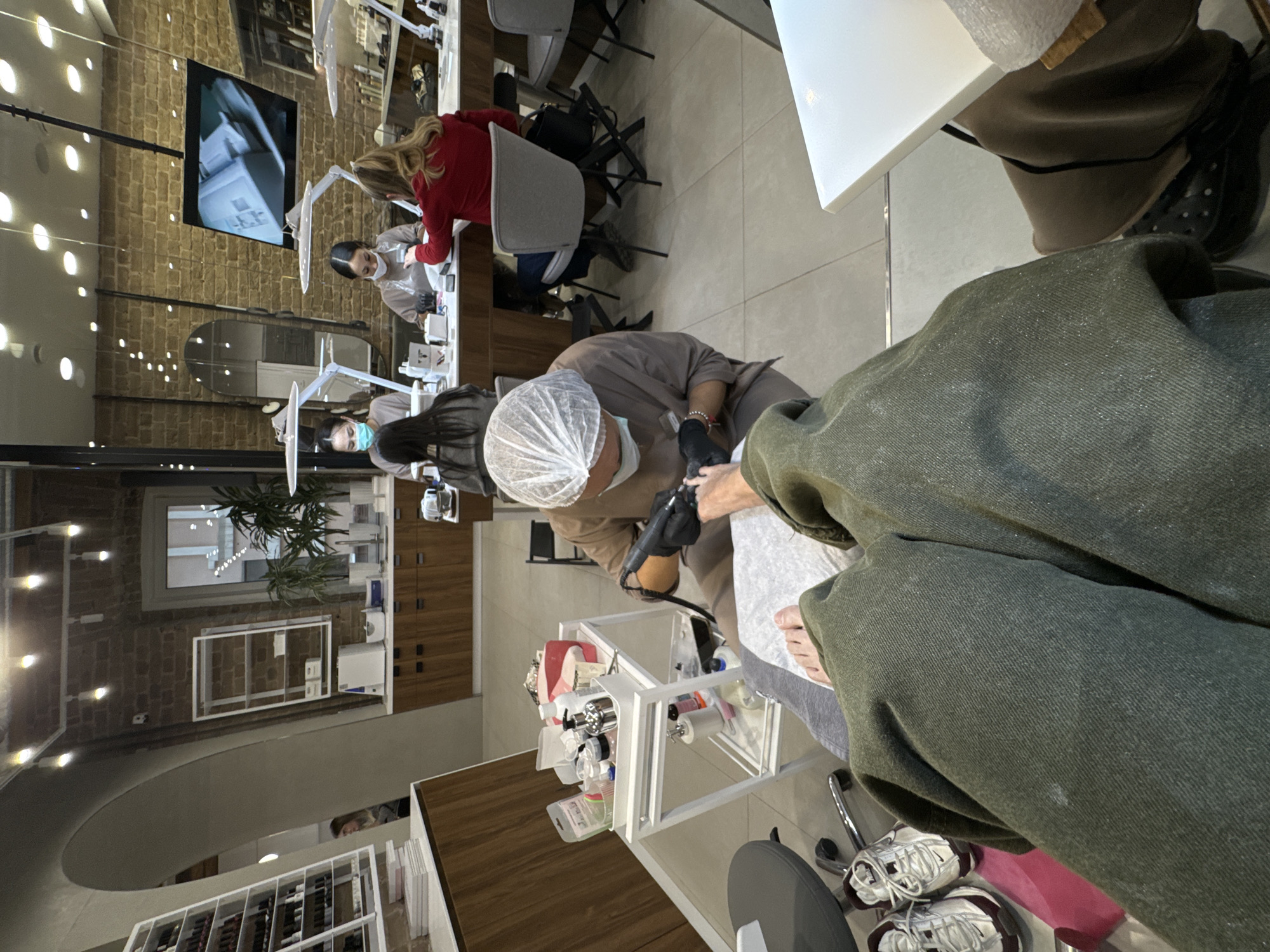 The image captures an interior view of a contemporary nail salon or beauty studio, taken from the perspective of a client receiving services.
In the immediate foreground, a person's bare foot is visible, resting on a white cushion. A nail technician, dressed in a brown uniform, a white hairnet, and black gloves, is focused on the foot, using an electric nail file or drill. A white, multi-tiered rolling cart, filled with numerous bottles of nail polish, lotions, and other manicure/pedicure tools, is positioned next to the technician. The client's leg, clad in olive green pants, is partially visible, and white sneakers with maroon accents are on the floor nearby.
The background reveals several other workstations. To the left, another technician, wearing a light-colored top and a face mask, is working on a client who is wearing a red top. Above this station, a flat-screen monitor displays a close-up image, likely related to nail care. Further back, another workstation with a technician is visible.
The salon's interior features a blend of textures, with exposed brick walls contrasting with smooth light-colored surfaces and rich wooden cabinetry. The floor is covered with light-colored tiles. The space is brightly lit by numerous recessed ceiling lights and adjustable task lamps positioned at each station. Decorative elements include a large mirror on the wall and potted plants, contributing to a professional and clean aesthetic. No discernible text is clearly visible on any products or signage.