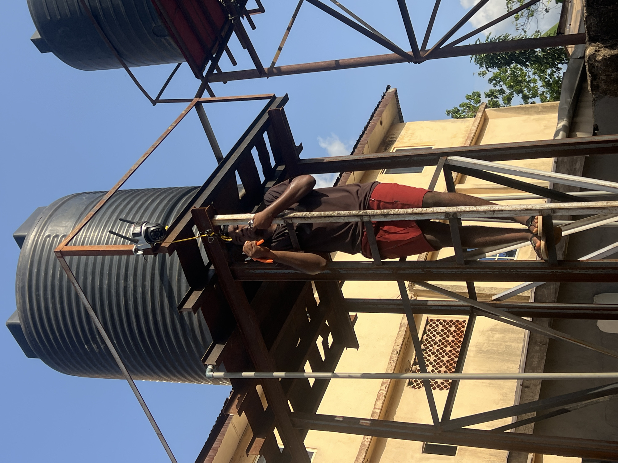 A man wearing a brown shirt and red shorts is working on a metal structure that supports two large black water tanks. He is holding a tool with orange handles. The structure is positioned on top of a yellow building with a patterned window. The sky is blue and partly cloudy. The location is Unknown, Unknown.