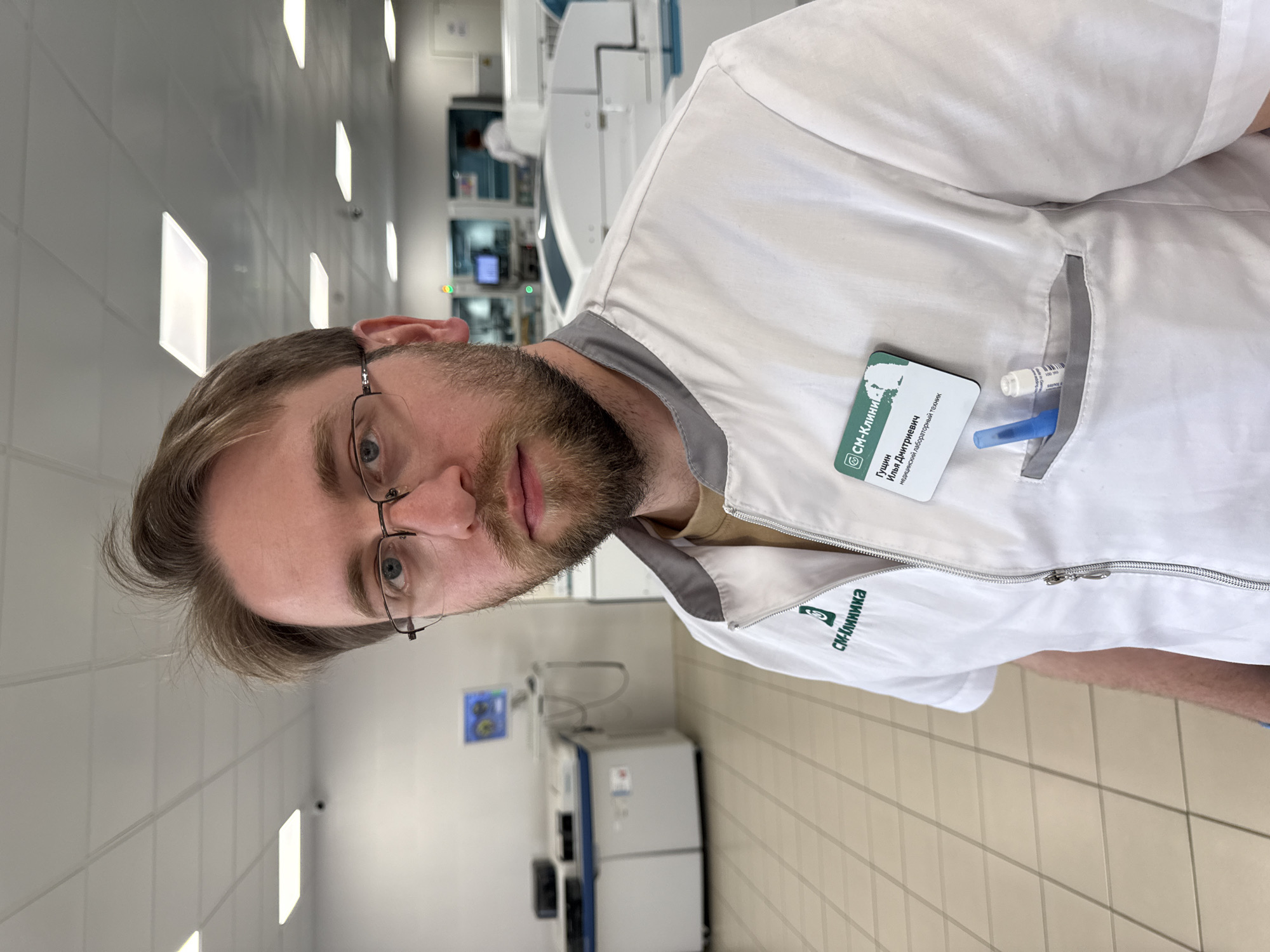 A man is centrally featured in a well-lit indoor laboratory setting in Moscow, Russia. He has short brown hair, a beard, and wears glasses, looking directly at the camera with a neutral expression. He is dressed in a white medical lab coat with grey trim and a zipper down the front.
Clipped to his chest pocket is a green and white name tag with the "SM-Клиника" (SM-Clinic) logo. The text on the tag identifies him as "Гущин Илья Дмитриевич" (Gushchin Ilya Dmitrievich) and states his profession as "медицинский лабораторный техник" (medical laboratory technician). A smaller embroidered "SM-Клиника" logo is also visible on the lower part of his lab coat. Inside his chest pocket, there appear to be a pen and a small vial.
The background reveals a clean, professional laboratory environment with white tiled walls and light-colored floor tiles. Several large, modern medical or analytical machines and equipment, some with screens and indicator lights, are visible, confirming the setting as a medical or research facility. The overall impression is that of a professional in his workplace, posing for a photo.