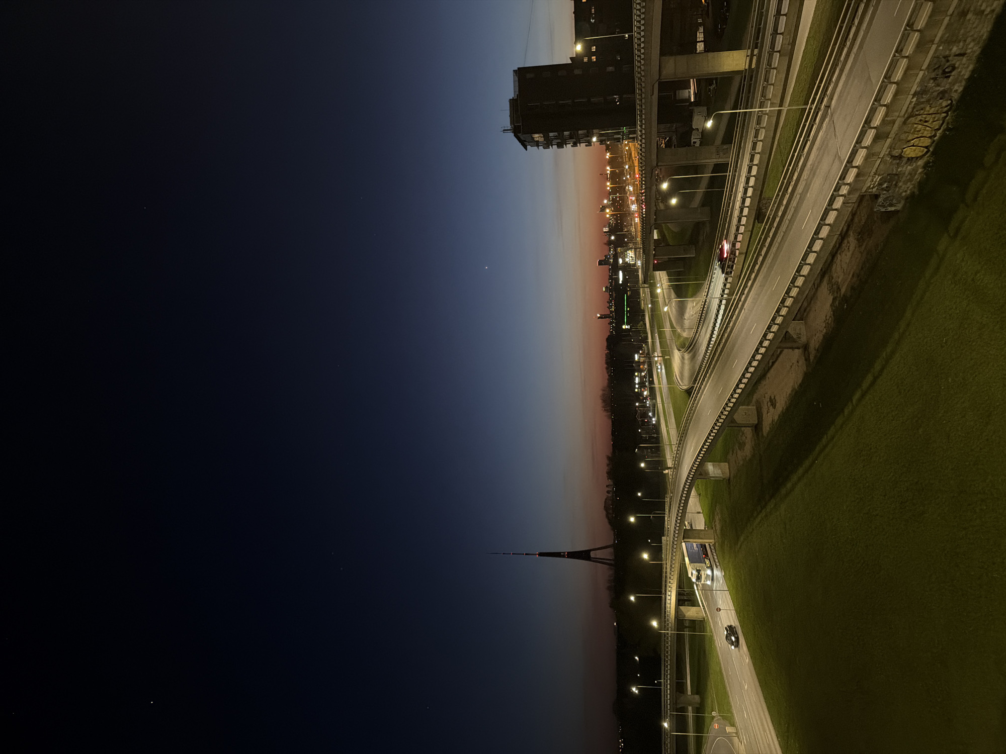 This image depicts a nighttime cityscape in Riga, Latvia, featuring the Riga Radio and TV Tower in the distance. The photo is taken from an elevated perspective, overlooking an overpass with traffic moving along it. Streetlights illuminate the roads and the adjacent grassy areas. In the background, there's a gradient in the sky from dark blue to lighter orange, with city buildings visible on the horizon. A building is visible from the top right corner.