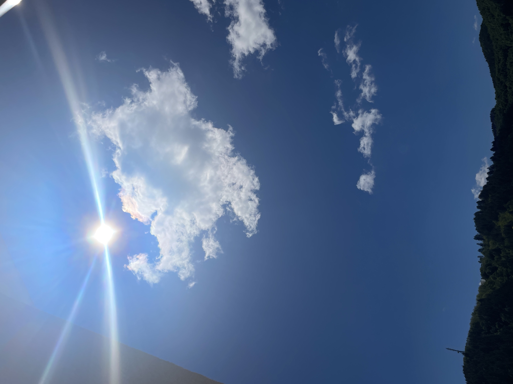 The image shows a bright, sunny day in Zlatograd, Bulgaria. The sky is a deep blue, with scattered white clouds. The sun is shining intensely, with bright rays emanating from it. Part of a building is visible in the lower-left corner. There is a tree line on the right side of the image.