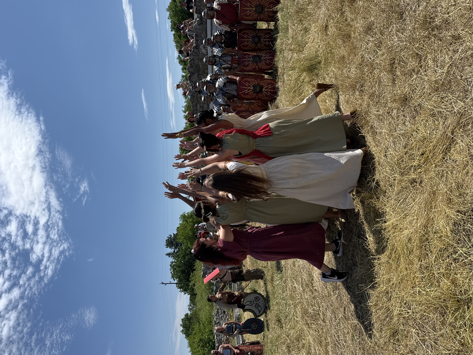 The image depicts an outdoor historical reenactment event under a clear blue sky with scattered white clouds, likely taking place at an archaeological site in Gigen, Bulgaria. In the foreground and midground, a circular formation of approximately seven women, dressed in long, flowing tunics or dresses in shades of maroon, olive green, and off-white, have their arms raised above their heads, forming a central focal point. The woman in the immediate foreground, positioned with her back partially to the viewer, wears a maroon dress and modern black athletic shoes with white soles. Other women wear flat, earthy-toned footwear. Their collective posture suggests a dance, ritual, or celebratory gesture.
To the left of the women, in the midground, are several male participants dressed as ancient warriors or gladiators. One figure stands prominently holding a large, round shield with a distinctive eye-like design and what appears to be a short sword. Another warrior carries a more generic round shield. Behind these figures and further to the left are visible stone ruins. In the midground to the right of the women, a line of Roman legionaries stands in formation, equipped with full segmented armor (lorica segmentata), distinctive red rectangular scutum shields adorned with golden emblems (possibly eagles or thunderbolts), and helmets. There are at least six legionaries visible.
In the background, behind the Roman soldiers and atop a stone wall structure, a group of spectators is observing the event. The ground throughout the scene is covered in dry, yellowed grass or straw. Further in the background, green trees and some electrical power lines are visible, stretching across the landscape. The sunlight suggests a clear, bright day, casting shadows on the dry ground.