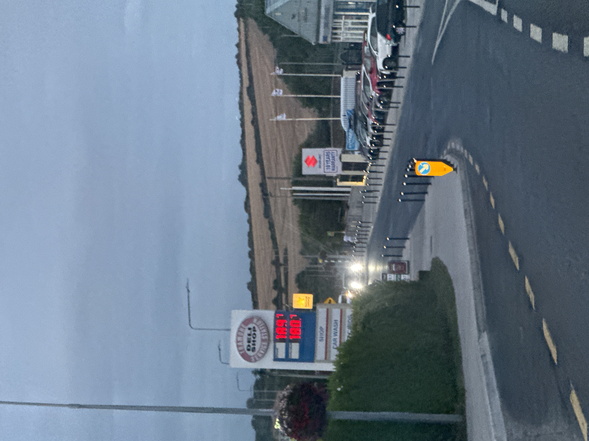The image depicts a street scene in Tramore, Ireland, at dusk. A prominent gas station sign displays fuel prices of 189.7 and 180.0. The gas station also features signs for a deli, shop, car wash, and credit cards. To the right, a Suzuki dealership is visible with several cars parked outside and a sign indicating a "10 YEARS WARRANTY." A row of bollards lines the street, and a yellow traffic cone with a blue symbol is placed on the pavement. The road is wet, suggesting recent rain, and headlights from oncoming vehicles are visible. In the background, rolling hills with dry vegetation suggest a rural setting bordering the town. The sky is overcast, contributing to the dim lighting conditions.
