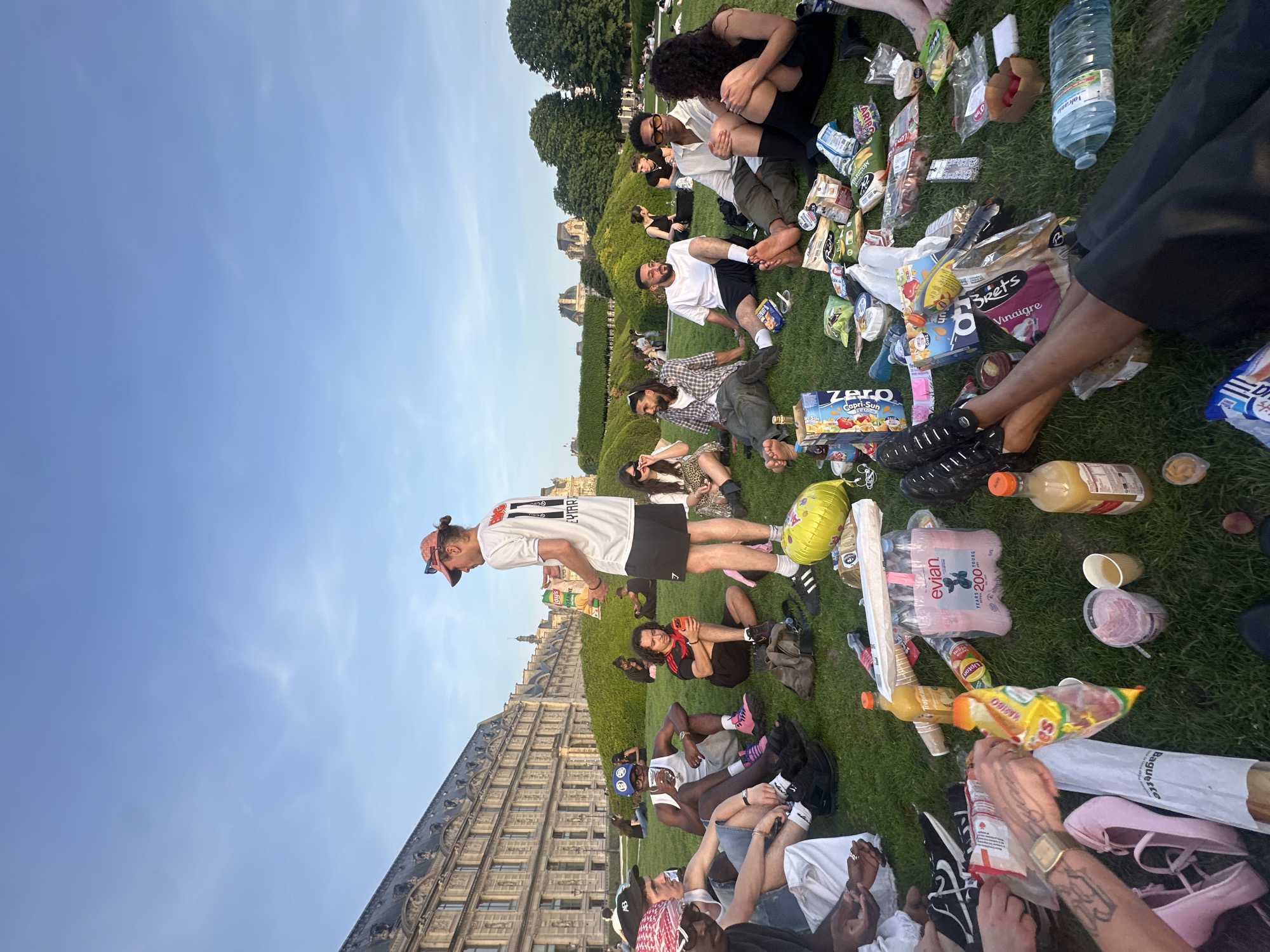 A group of people are gathered on a grassy lawn littered with snacks and drinks. The group appears to be in a park with a building with many windows in the background. Many people are seated or lying down in the grass. A man in a soccer jersey is standing in the center of the lawn. The setting suggests a relaxed, informal gathering in a park. In the background is the Louvre Museum in Paris.