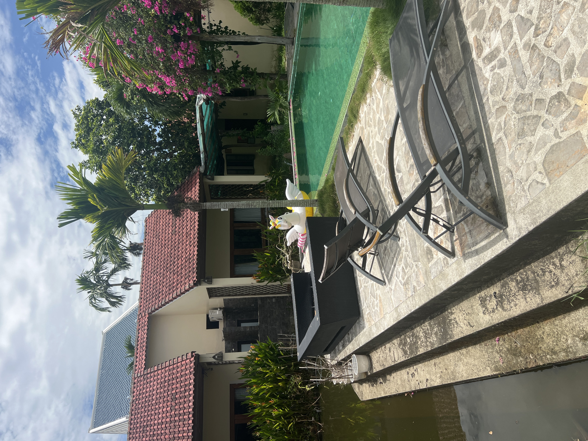 The image depicts a residential property with a swimming pool, likely in Hoi An, Vietnam, given the architectural style of the houses and the presence of tropical vegetation. In the foreground, two dark grey lounge chairs are positioned on a stone patio. Behind the chairs, a large, dark woven rectangular planter box sits on a raised concrete platform, adjacent to a body of water on the left. Further back, a bright yellow inflatable unicorn floats in the turquoise water of the rectangular swimming pool. The pool is bordered by a green tiled edge. Behind the pool, lush green trees and vibrant pink bougainvillea flowers are visible, with palm trees also present. The houses feature red-tiled roofs and cream-colored walls. The sky above is partly cloudy with white cumulus clouds. The overall impression is a serene and tropical outdoor living space.