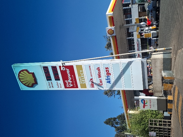 The image shows a Shell gas station in Gitaru, Kenya, located at latitude -1.226 and longitude 36.664. The gas station is open and operational. There are several vehicles parked, and several people are present, including some working at the gas station. The sign indicates that the station sells fuels unleaded, fuel-flow diesel, and Shell V-Power performance fuel. The sign also indicates that the station offers Shell Select and a car wash. The gas station is located in a rural setting with trees in the background. The sky is blue and cloudless. The signage features red and yellow details. The station has a concrete base and a large canopy covering the fuel pumps. An Afrigas sign is visible below the Shell Select sign. There is a green, rectangular sign with a white logo on it. There are also yellow and black striped markings on the pavement near the sign. There is a fence on the left side of the image.