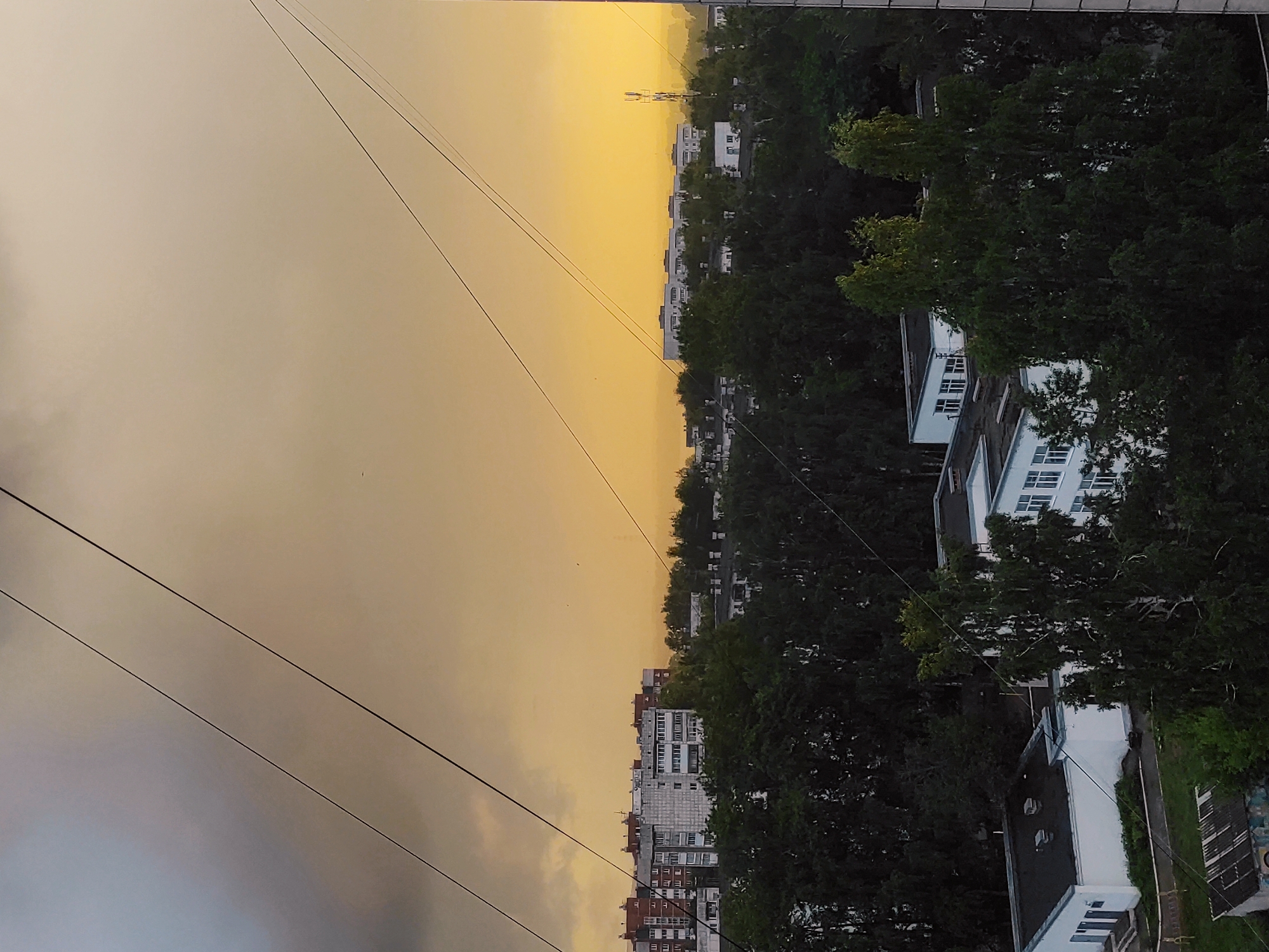This media file captures a panoramic view of an urban landscape, likely at sunrise or sunset, given the striking golden-yellow hue of the sky on one side. The image, originally rotated, depicts a horizontal horizon line separating the sky from a city below.
The sky transitions from a vibrant, warm golden-yellow on the right to a softer, hazy grey on the left, suggesting either atmospheric conditions or an approaching weather front. Several dark, thin power lines or utility cables cut diagonally across the sky from the upper left to the lower right.
Below the horizon, a densely built urban area unfolds. Numerous multi-story buildings, varying in color from white and grey to reddish-brown, line the cityscape. Many of these structures appear to be residential, featuring multiple windows. The buildings are interspersed with and often partially obscured by abundant, dark green trees and dense foliage, indicating a well-treed environment. In the distance, against the bright yellow sky, a tall, slender structure resembling a cell tower or antenna stands prominently.
No specific human activity is visible, and the scene appears tranquil, dominated by the interplay of natural light and urban architecture. There is no visible text in the image. The setting is consistent with an urban or suburban area, possibly within Chaikovsky, Russia, given the provided context.