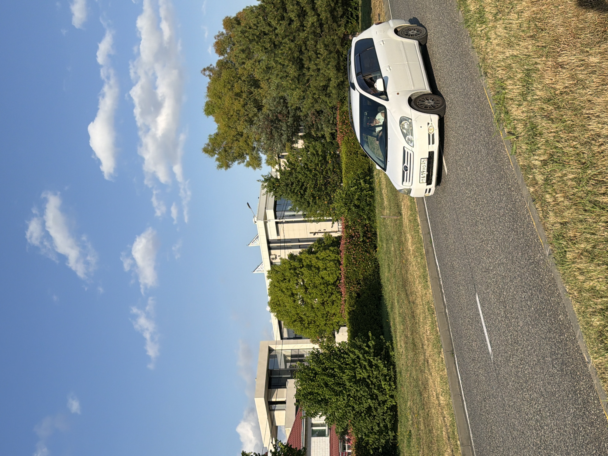 A white minivan is parked on the right side of a paved road, facing away from the viewer. The minivan is a tall, compact vehicle with tinted windows. A person, likely the driver, is visible through the driver's side window, wearing a light-colored shirt. The road has a white line marking its center. To the right of the road, there is a strip of dry, yellowed grass. To the left of the road, there is a line of green bushes and trees, with modern-looking buildings visible behind them. The buildings are made of light-colored materials and have large windows. A bright blue sky with scattered white clouds is visible above the trees and buildings. The overall scene suggests a daytime setting, possibly late afternoon due to the angle of the light. The license plate on the front of the minivan is visible and appears to read "T619YO24". The location is identified as Sirius, Russia.