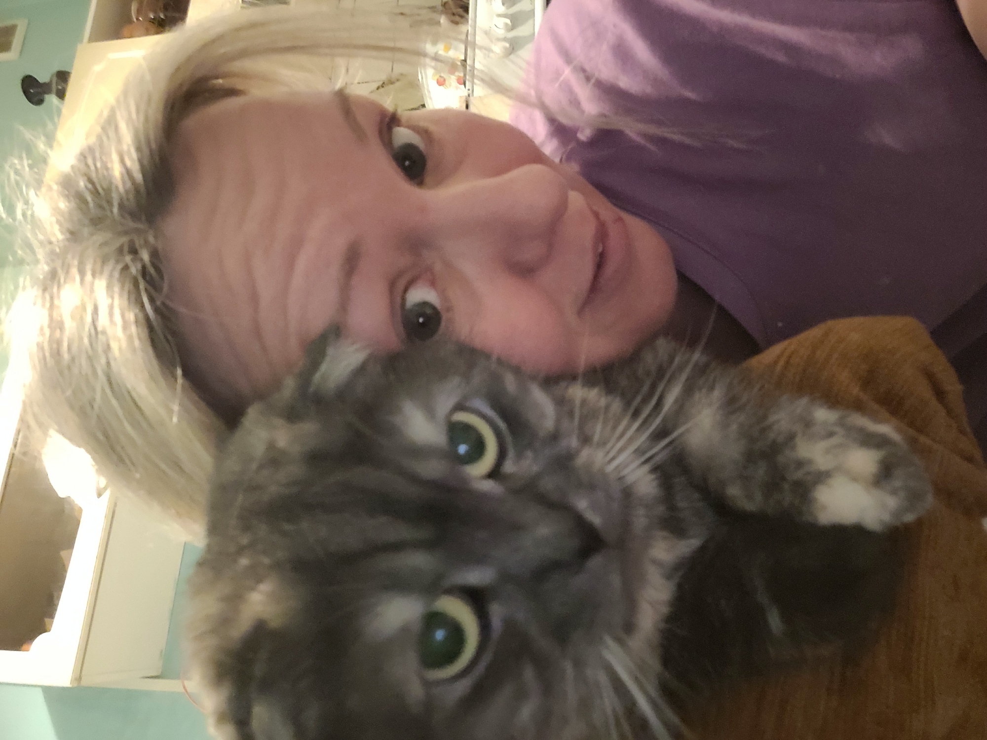 A woman with blonde hair and a purple shirt is holding a gray and white cat. The cat has large green eyes and a white spot on one of its paws. The background is blurry but shows a white cabinet and light blue wall. There is no indication of the location being Izhevsk, Russia.