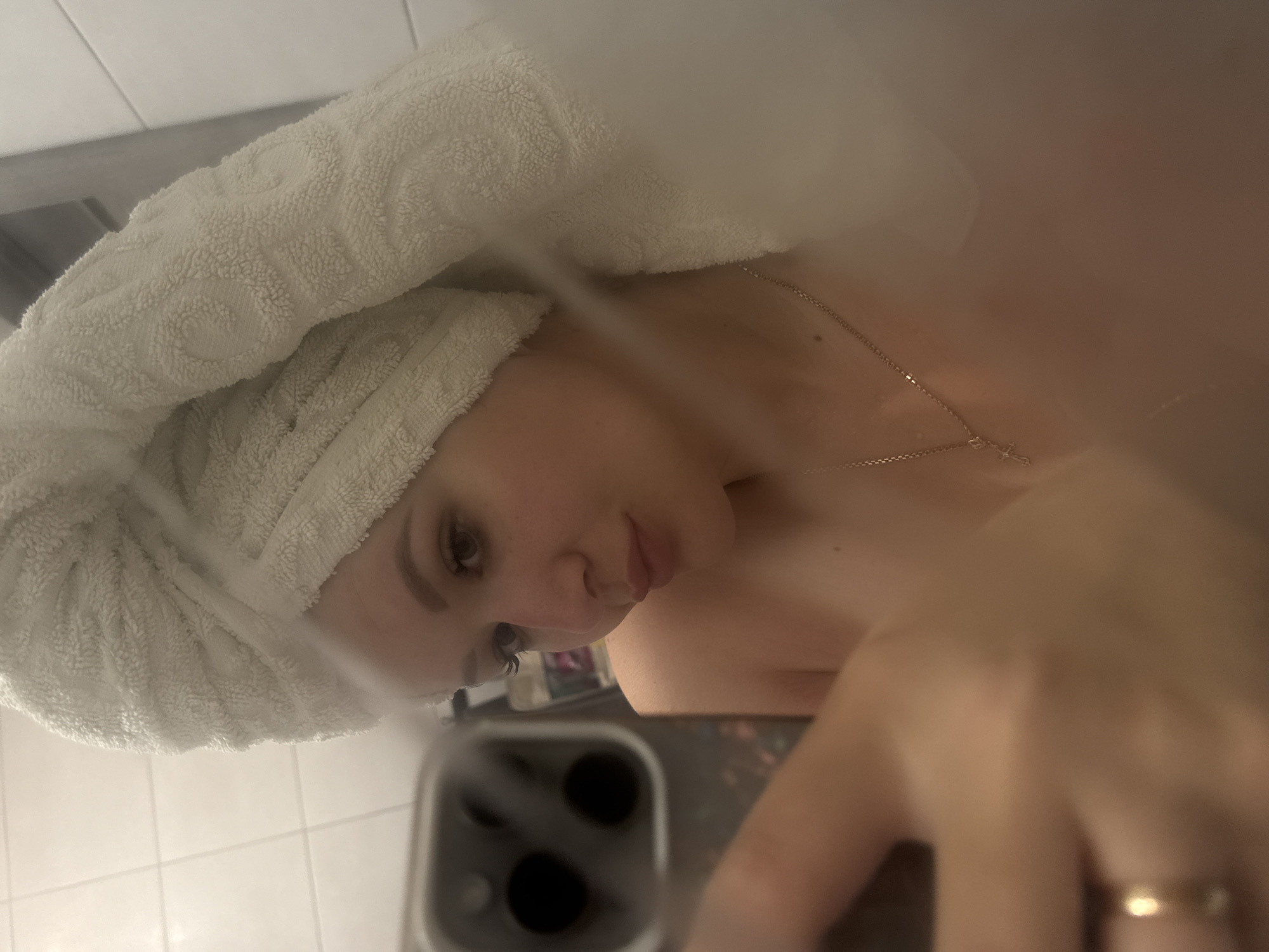 A woman is taking a selfie in a bathroom. She has a white towel wrapped around her head and is holding a smartphone in her right hand. She is wearing a delicate gold necklace. The bathroom has white tiles. The location is unspecified; there is no visible evidence to suggest it is Gelendzhik, Russia.