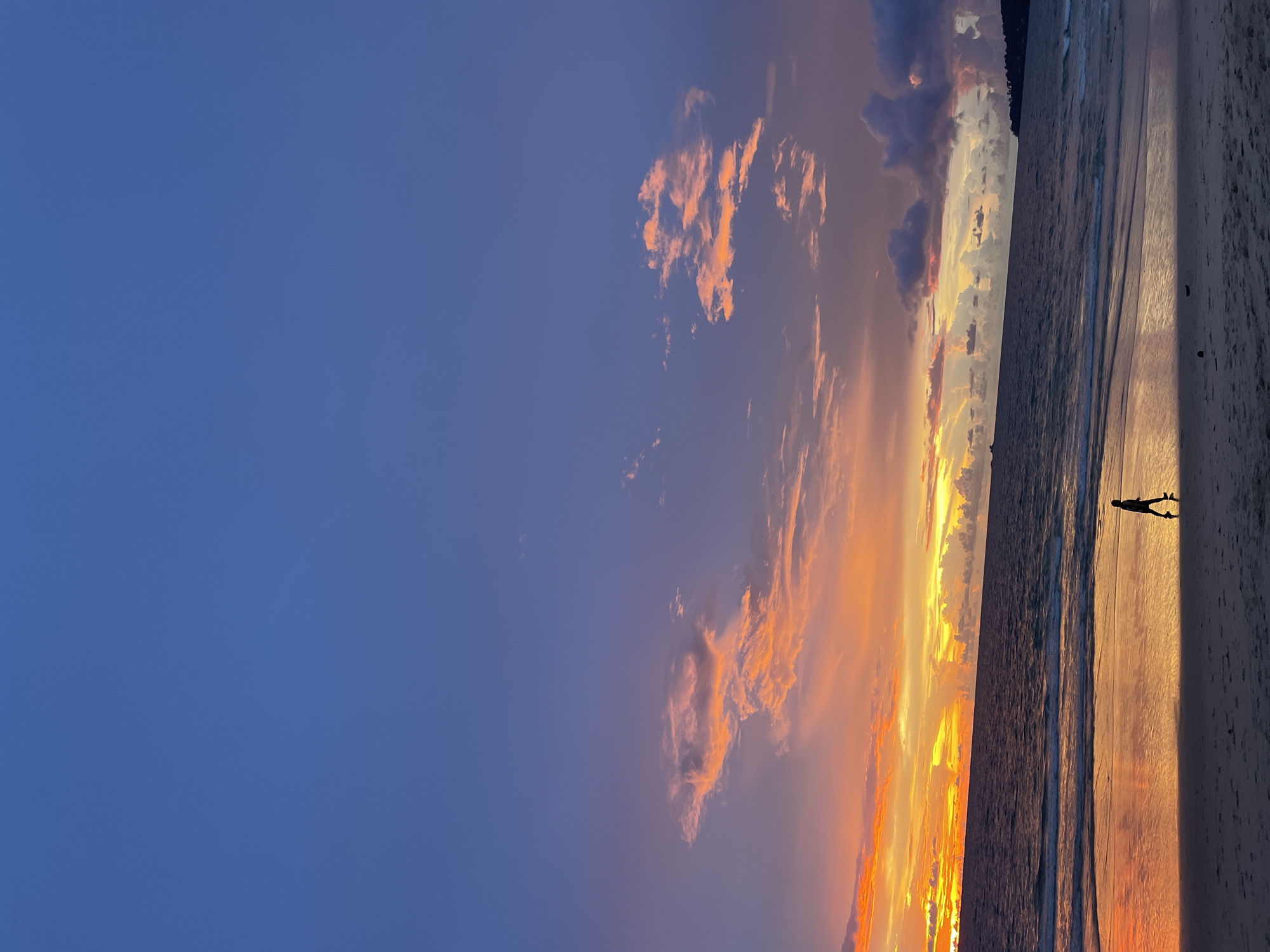 A solitary figure walks along a sandy beach in Choeng Thale, Thailand, during a vibrant sunset. The sky is a gradient of deep blue at the top, transitioning to hues of purple, orange, and yellow near the horizon, where the sun has just set. Fluffy clouds are illuminated by the setting sun, casting fiery colors across the sky. The calm ocean reflects the colorful sky, creating a mirror-like effect on the wet sand. The beach is mostly empty, with only the silhouette of the person walking. The overall scene evokes a sense of peace and natural beauty.