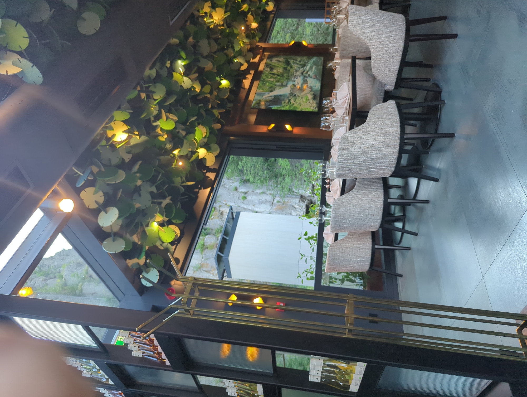 The image depicts an interior scene of a restaurant with a contemporary design. The setting appears to be a dining area with multiple tables and chairs. The tables are set with glassware and napkins, suggesting a prepared meal or service is imminent. The chairs are upholstered in a neutral, textured fabric and have dark wooden legs.
A prominent feature of the restaurant's decor is a ceiling installation resembling lily pads, adorned with small, warm-toned lights. This artistic element creates a unique and possibly whimsical ambiance. Large windows offer glimpses of the outdoors, showing trees and a rocky landscape, hinting at a natural or scenic location. The lighting in the restaurant is a mix of the decorative lily pad lights and ambient overhead lighting, casting a warm glow.
There are no people visibly present in the image, suggesting the restaurant is either closed, empty at the moment, or the photo was taken before or after operating hours. The overall impression is one of a stylish and possibly upscale dining establishment, focusing on atmosphere and aesthetics. The time of day is difficult to ascertain definitively, but the warm indoor lighting might suggest evening or a dimly lit interior. There is no visible text in the image.