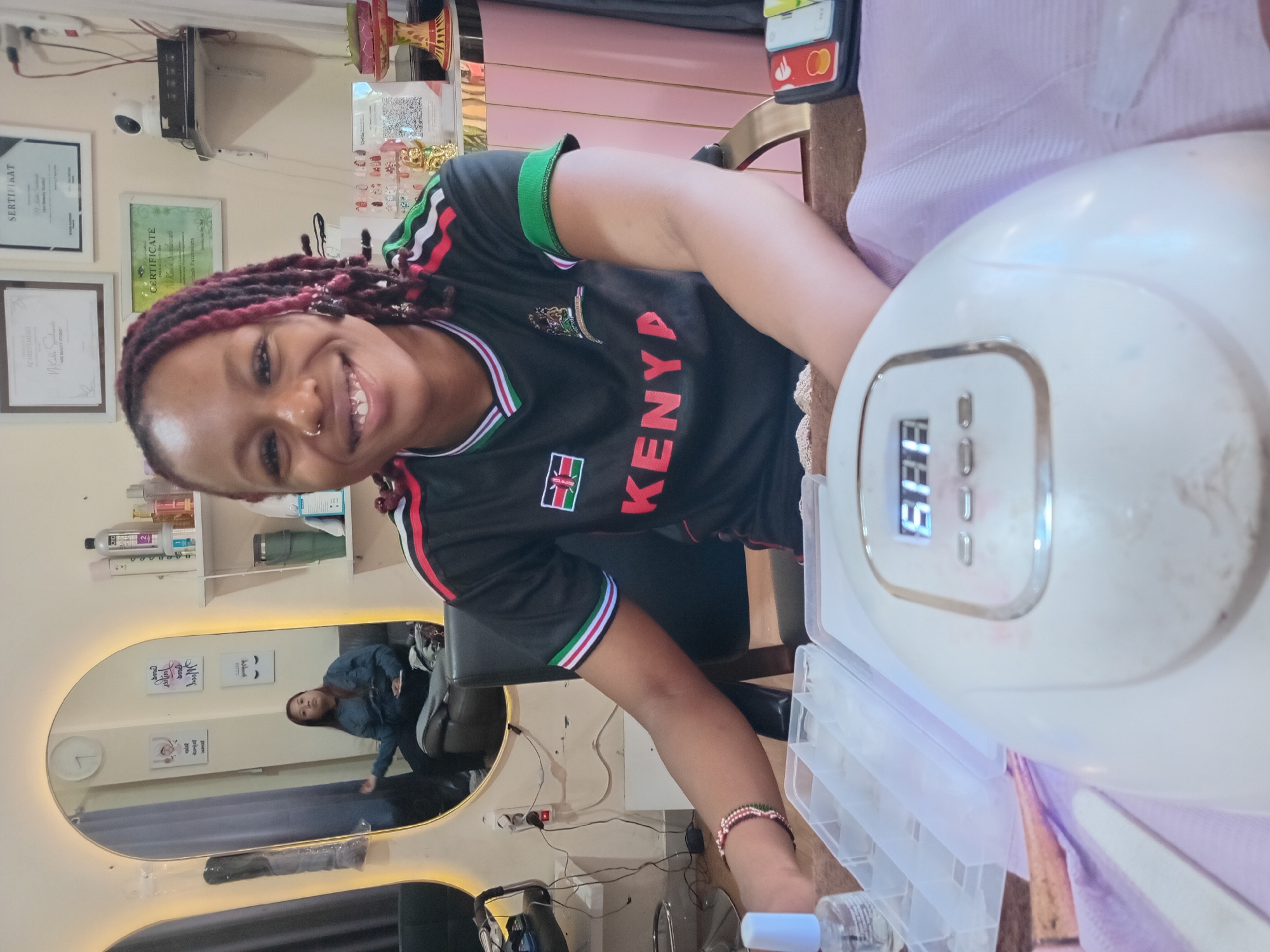 This image captures a vibrant scene inside a beauty salon or nail bar in North Kuta, Indonesia. The main subject is a smiling woman with dreadlocks and a nose piercing, wearing a black t-shirt adorned with "KENYA" and the Kenyan flag. She is positioned to the right side of the frame, looking directly at the camera with a cheerful expression, suggesting a friendly interaction or photo opportunity.
Her hands are near a white nail drying lamp, which displays "588" on its digital screen, possibly indicating a timer setting. Next to the lamp is a clear plastic organizer tray, likely holding nail art supplies or tips.
The background reveals more details of the salon: framed certificates are visible on a light-colored wall, including ones labeled "SERTIFIKAT," "CERTIFICATE," and "ACHIEVEMENT," with "French Technique" also partially legible on one. A security camera is mounted high on the wall. Shelves stocked with various beauty products can be seen. A large, oval mirror with illuminated edges reflects another person, likely a client, seated in a salon chair in the background. A small phone holder displaying logos resembling credit cards (e.g., Mastercard) is also visible near the lamp.
The overall atmosphere appears professional and welcoming. The image is taken at a slightly tilted angle.