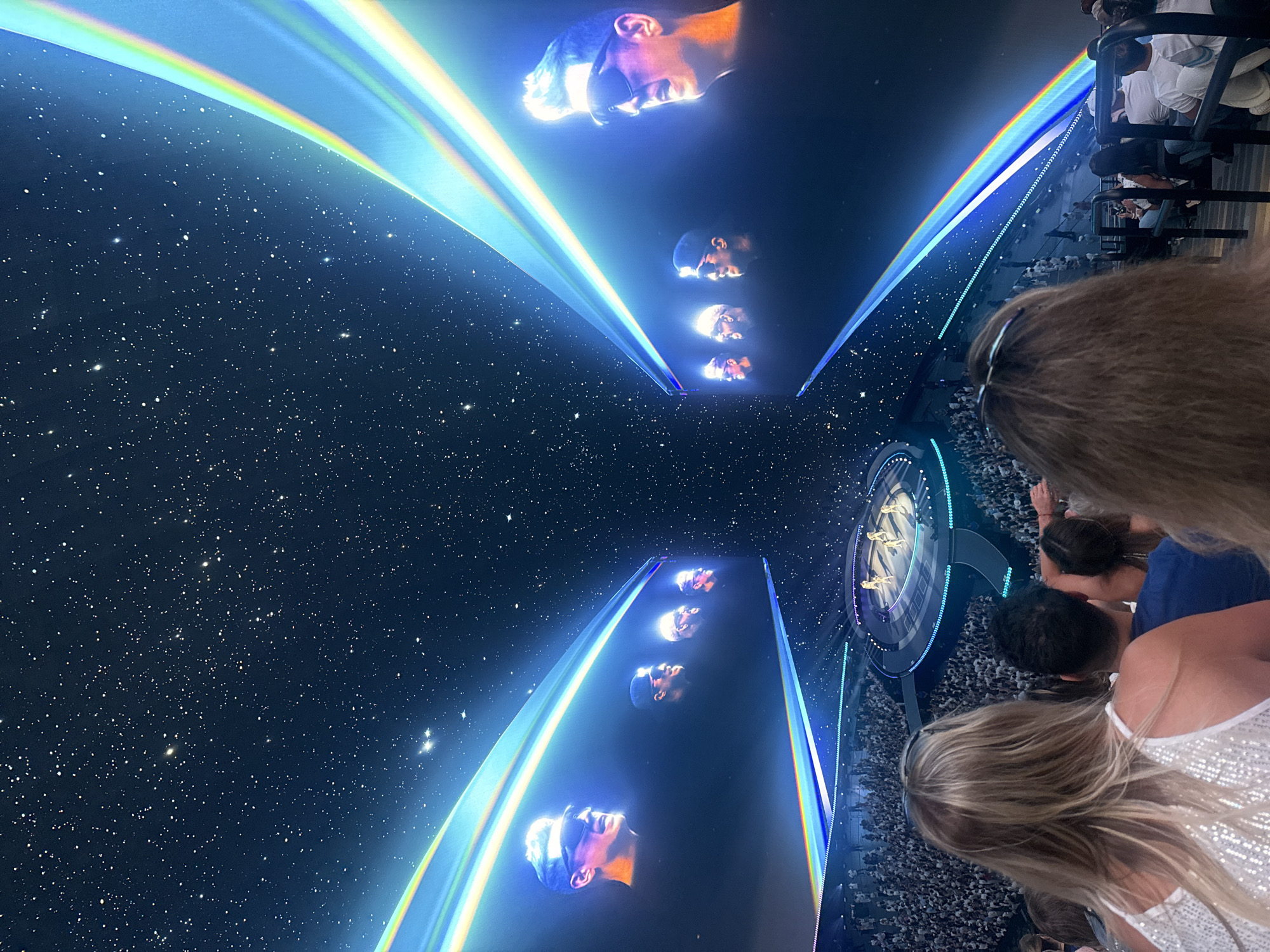 This image depicts a concert or performance taking place in the city of Paradise, United States of America. The scene is dominated by a large, immersive projection of a starry night sky, with diagonal, rainbow-colored light beams extending from the sides. On large screens on either side of the venue, close-ups of four men wearing sunglasses are displayed. A circular stage with performers is visible in the distance, surrounded by a large audience. In the foreground, the backs of several concert-goers are visible, with their hair and shoulders in frame. The overall atmosphere suggests a technologically advanced and visually striking entertainment event.