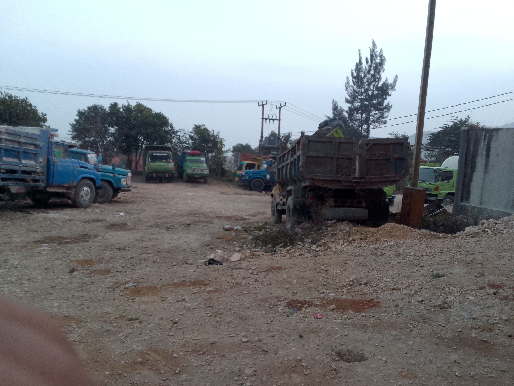 The image depicts a lot with multiple trucks parked on a dirt and gravel surface, likely in an industrial or construction area in Gunungmasigit, Indonesia. The sky is overcast, suggesting a cloudy day. Several large trucks, some with open beds carrying loads, are visible. These include blue, green, and a brown/red truck in the foreground. Trees are present in the background, along with utility poles and power lines. A concrete structure, possibly a wall or building, is on the right side of the image. The ground is uneven and scattered with small rocks and debris. No people are clearly visible in the image. The scene appears to be a storage or staging area for heavy vehicles.