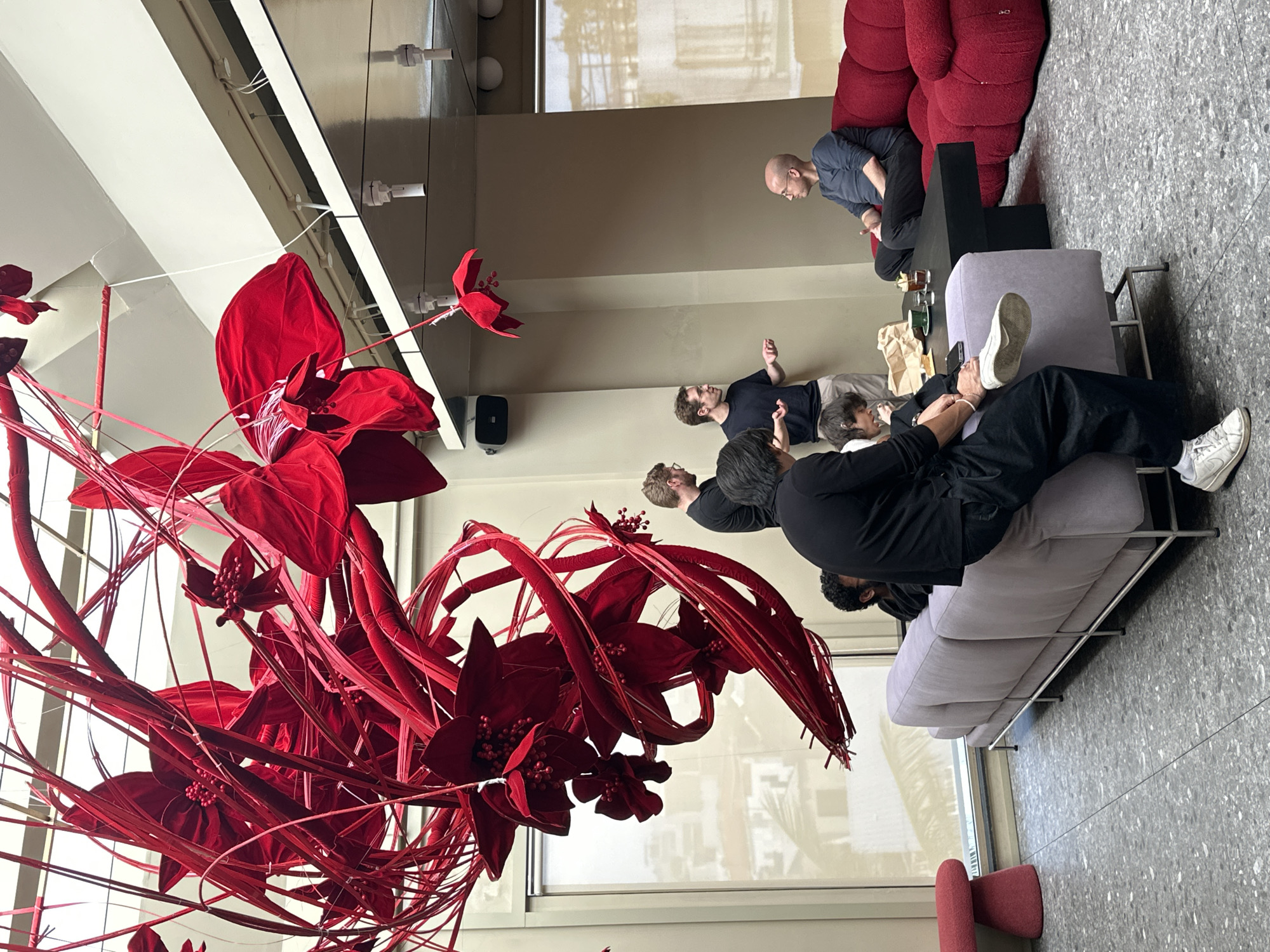 This image captures an indoor scene, possibly a lounge area or event space, characterized by a large, vibrant red floral art installation suspended from the ceiling. The art piece is intricate, with numerous red fabric elements resembling petals and tendrils, creating a dramatic and visually striking centerpiece.
In the foreground and midground, several individuals are present. On the left, a person wearing black pants and white sneakers is seated on a light purple couch, attending to someone's foot. To their left, a red stool with a rounded top is visible near a large window.
Several other individuals are interacting in the midground. Two men in black shirts are standing and conversing, one gesturing with his hands. Another man with glasses is also standing nearby, observing. Further to the right, a bald man in a dark blue shirt is seated on a plush red sofa, facing the group. A dark coffee table sits between the sofas, holding drinks and what appears to be a paper bag.
The environment features light-colored walls and a textured, speckled floor. The lighting suggests an interior space with natural light filtering through the windows. The overall aesthetic appears modern and stylish, potentially a contemporary art gallery, a hotel lobby, or a public event space. The image does not provide any specific visual cues or landmarks to place it within the city of Đà Nẵng, Vietnam.