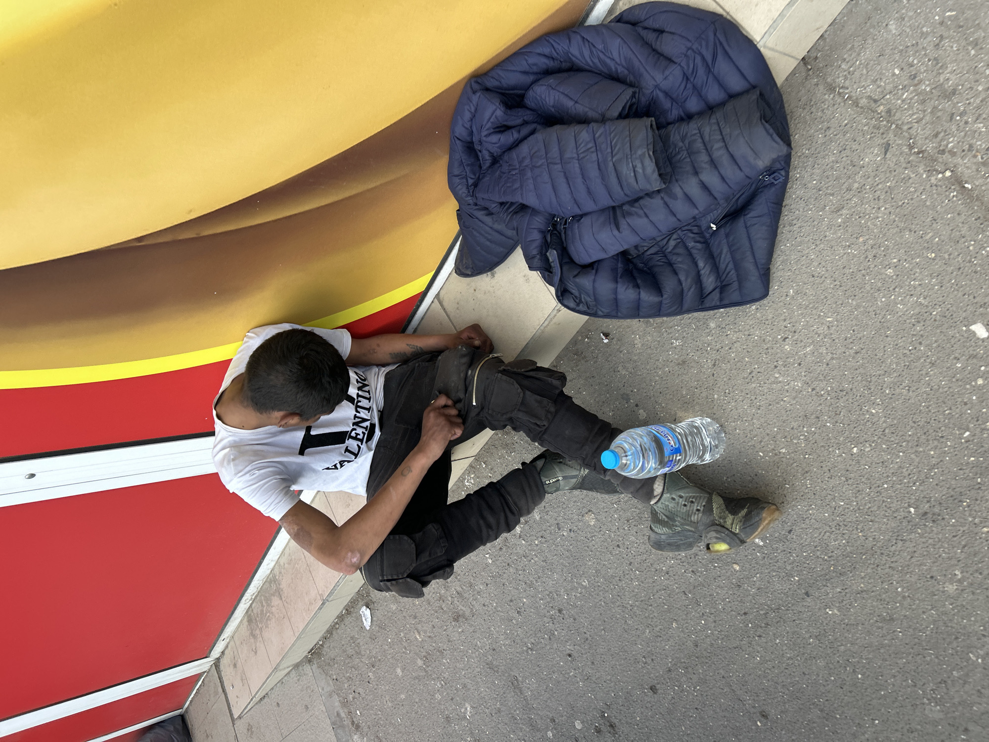 The image shows a man sitting on the sidewalk in Sofia, Bulgaria. He is leaning against a red and yellow wall. He is wearing a white t-shirt with "VALENTINO" printed on it, black pants, and dirty green sneakers. A bottle of water rests on his left leg. A blue, quilted jacket lies next to him. The sidewalk is gray concrete.