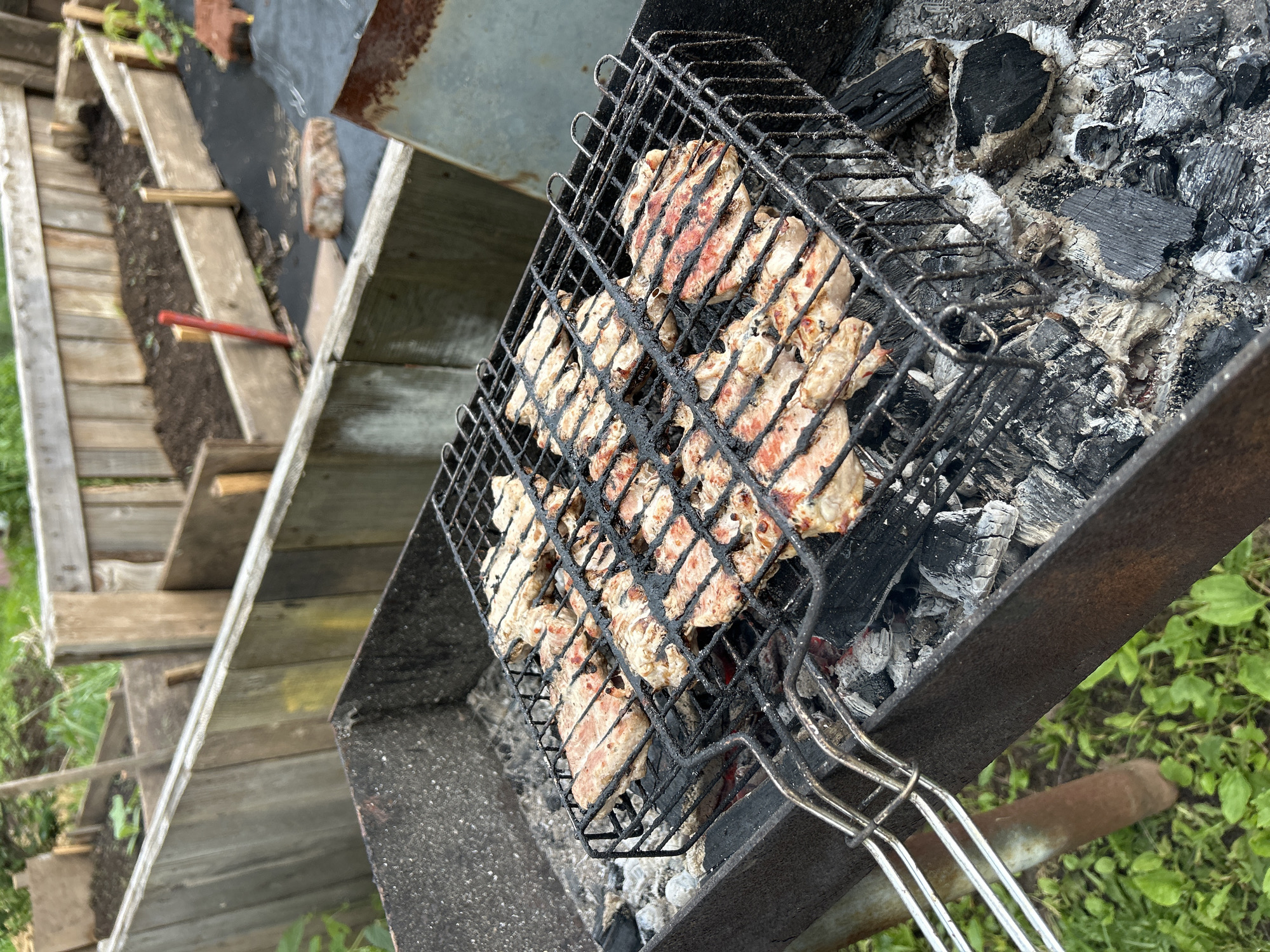 A barbecue grill filled with cooking meat sits outdoors on a grassy area. The grill is a black metal box with a wire grate holding several pieces of raw, seasoned meat, possibly chicken or pork. The meat shows visible grill marks and some pinkness, indicating it is in the early stages of cooking. Coals and ash are visible below the grate, with some embers glowing red. In the background, a wooden fence surrounds raised garden beds filled with dark soil. The overall setting suggests a casual outdoor meal preparation in a backyard or garden. The image does not contain any individuals or any contextual clues that would indicate the location as Азинский, Russia.