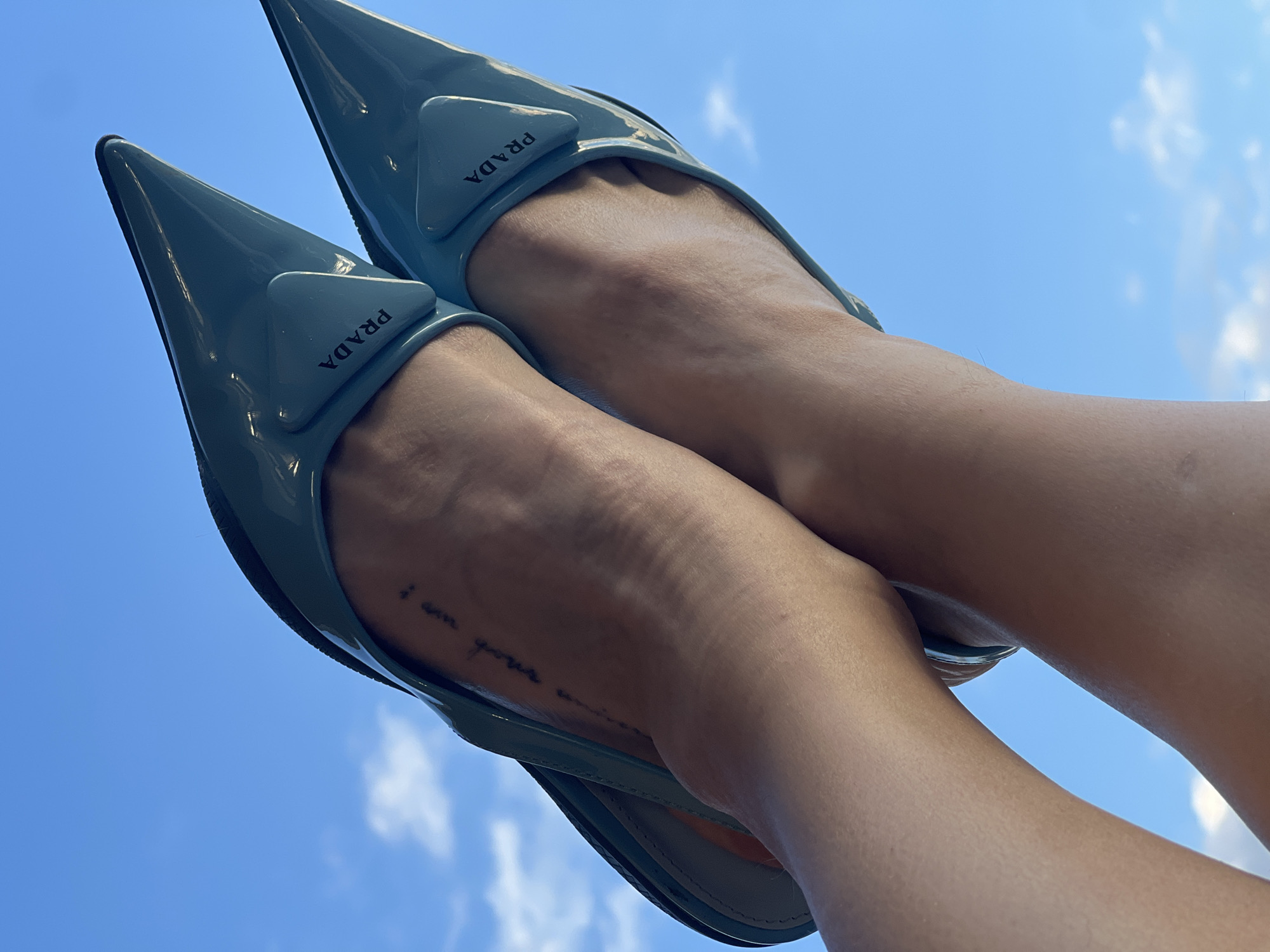 In a close-up, low-angle shot, a pair of light blue patent leather Prada mules with sharp pointed toes are worn by a person whose legs and feet are extended against a bright blue sky with scattered white clouds. The mules feature a triangular detail on the vamp with the Prada logo embossed in black. A small, faint tattoo is visible on the instep of the left foot. The background is a clear sky, suggesting an outdoor setting, possibly a rooftop or a balcony, in Saint Petersburg, Russia, though no specific landmarks are identifiable. The lighting suggests daytime.