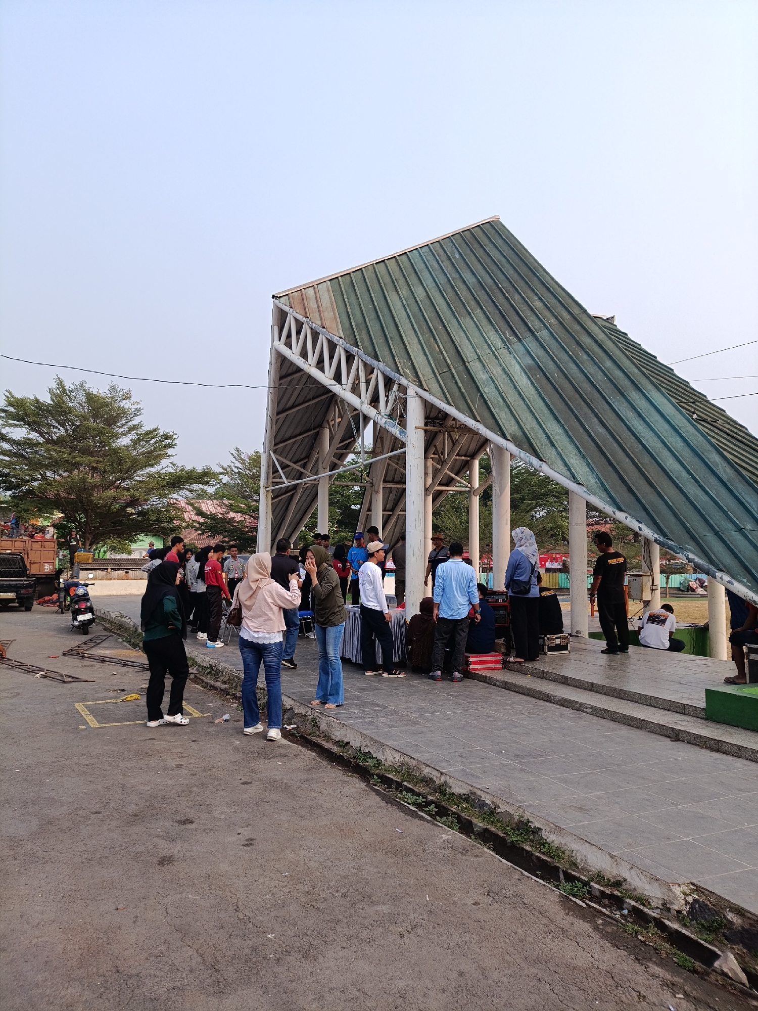 The image depicts an outdoor scene in the city of Cariu, Indonesia, likely at an event or gathering. A prominent feature is a modern-looking covered walkway or pavilion with a slanted, multi-paneled green roof supported by white pillars. The ground consists of a paved walkway and an asphalted area, with a concrete drainage ditch running along the edge of the walkway.
A diverse group of people, estimated to be around 15-20 individuals, are present. They are standing, sitting, and interacting in various small groups. Some individuals appear to be taking photos with their phones, while others are engaged in conversation. Clothing styles suggest a casual atmosphere, with a mix of long-sleeved shirts, t-shirts, jeans, and headscarves.
In the background, trees with dense foliage are visible, along with buildings and what appears to be a truck loaded with goods. A motorcycle is parked on the asphalted area. The sky is overcast or hazy, indicating diffused lighting. The overall impression is of a public space being utilized for a social event or meeting.
