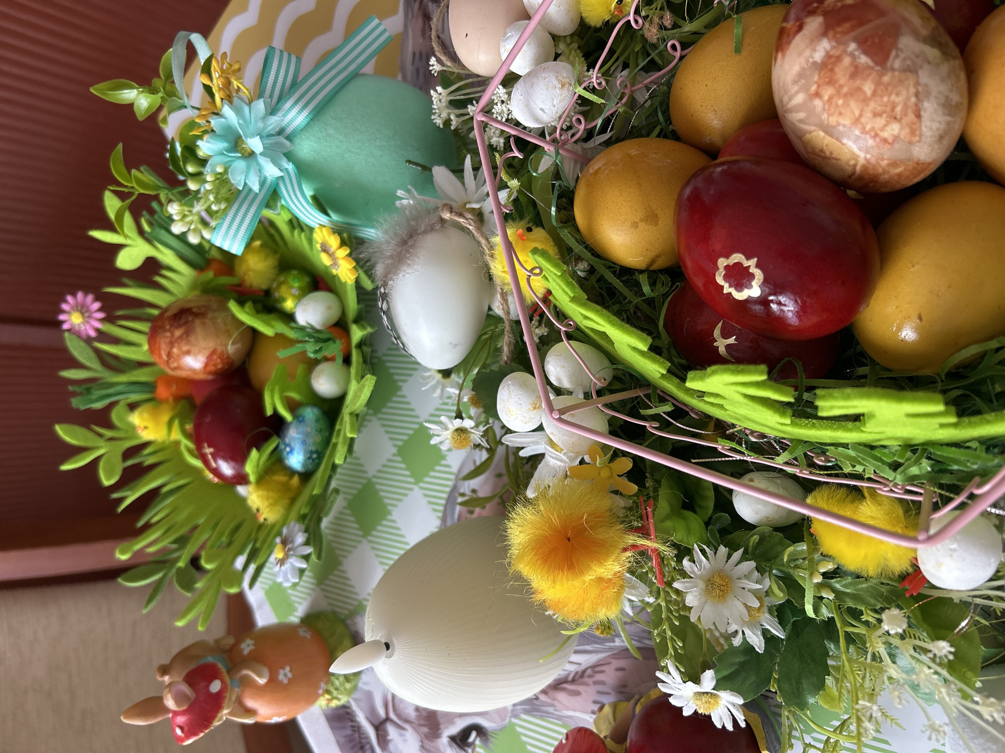 The image depicts a close-up of Easter decorations, likely indoors, but the specific location in Plovdiv, Bulgaria is not discernible from the image. The decorations are arranged as wreaths or centerpieces, featuring colored Easter eggs in various shades of red, brown, and yellow. There are also white eggs, some speckled, others decorated with feathers. Small yellow chick figures are nestled among the eggs and artificial green foliage. Daisies are interspersed throughout the decorations. Some of the eggs are adorned with small decorative elements. One wreath features a light blue bow. In the lower left corner, there is a small bunny figurine. The decorations are placed on a checkered surface, which might be a tablecloth or placemat.